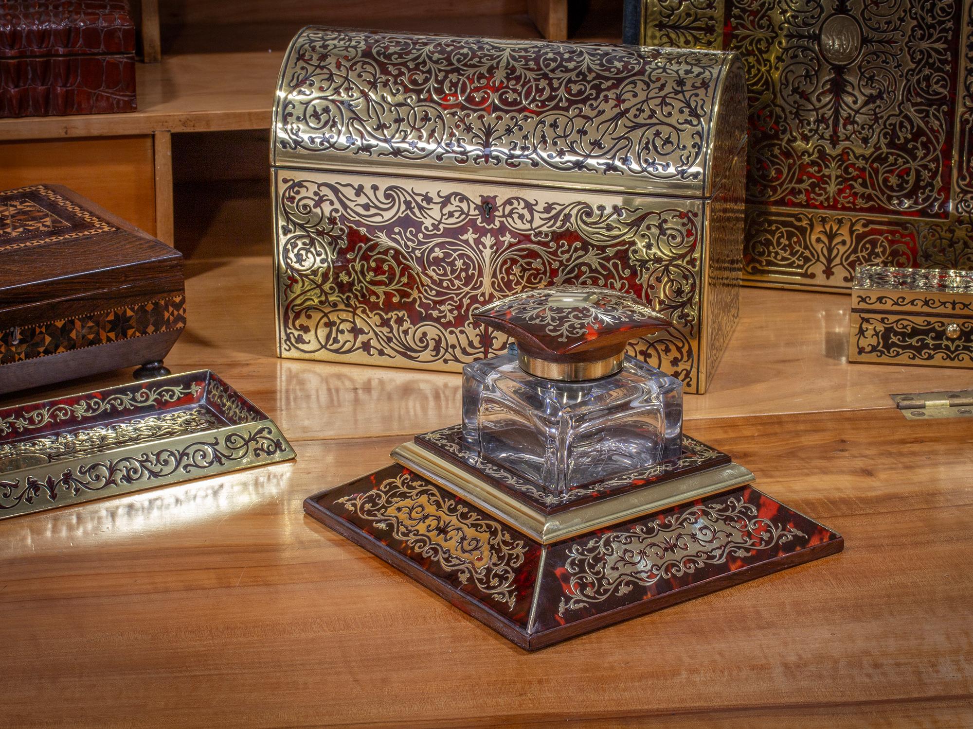 The inkwell with a square base having four faceted faces leading up to a central boulle lidded square glass inkwell with brass collar. The Boulle work finished in typical fashion with Tortoiseshell and Brass inlay. Signed to the centre of the stand