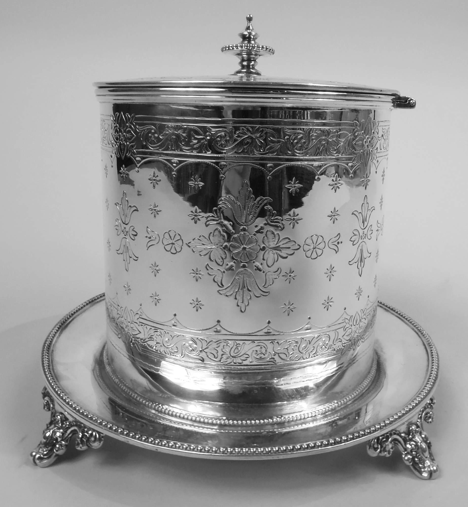 Victorian Classical sterling silver biscuit jar. Made by Henry Holland in London in 1872. Drum form; cover flat and hinged with vasiform finial. Engraved stylized floral ornament and oval frame (vacant). Mounted to round stand with four