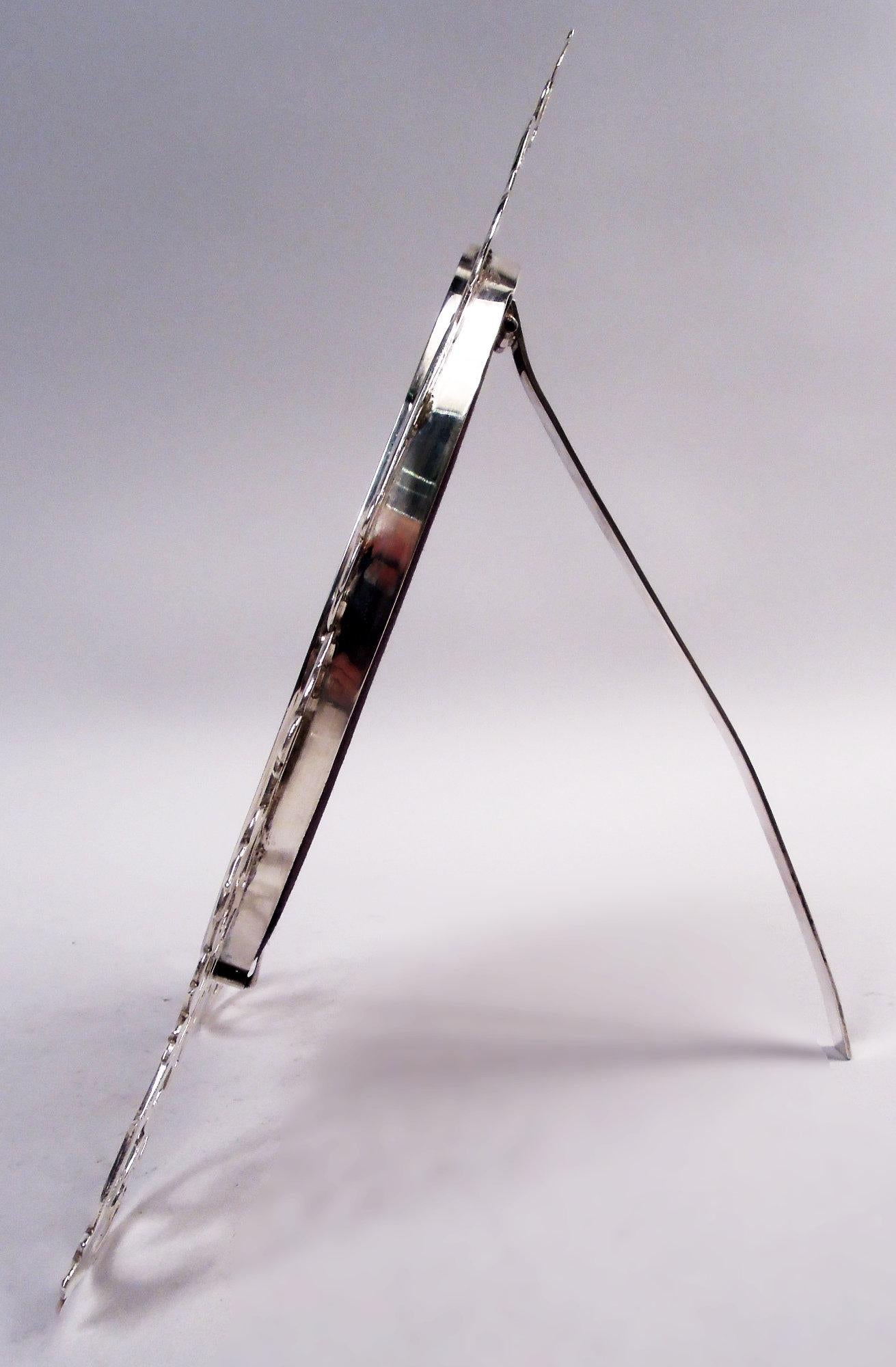 Victorian Classical sterling silver picture frame. Made by John Tiley in London in 1898. Plain and flat oval surrounded by open leafing scrollwork; at top solid oval (vacant). With glass, silk lining, and hinged easel silver support. Fully marked.