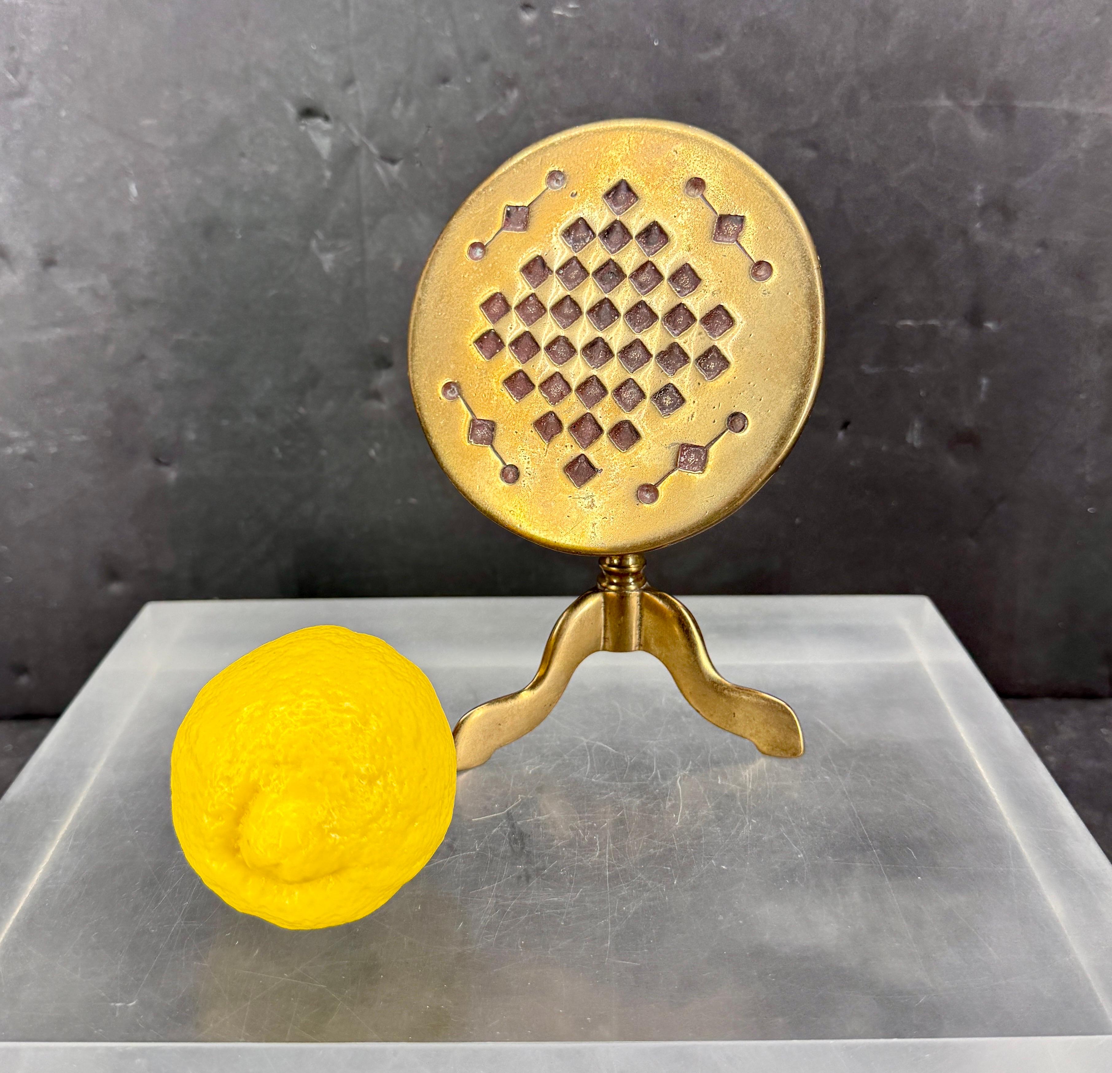 Encantadora mesa basculante de latón en miniatura, también conocida como reflector de velas. Es una pieza decorativa que presenta una parte superior redonda con un dibujo de tablero de ajedrez. 
La mesa se describe a menudo como una mesa de juego en