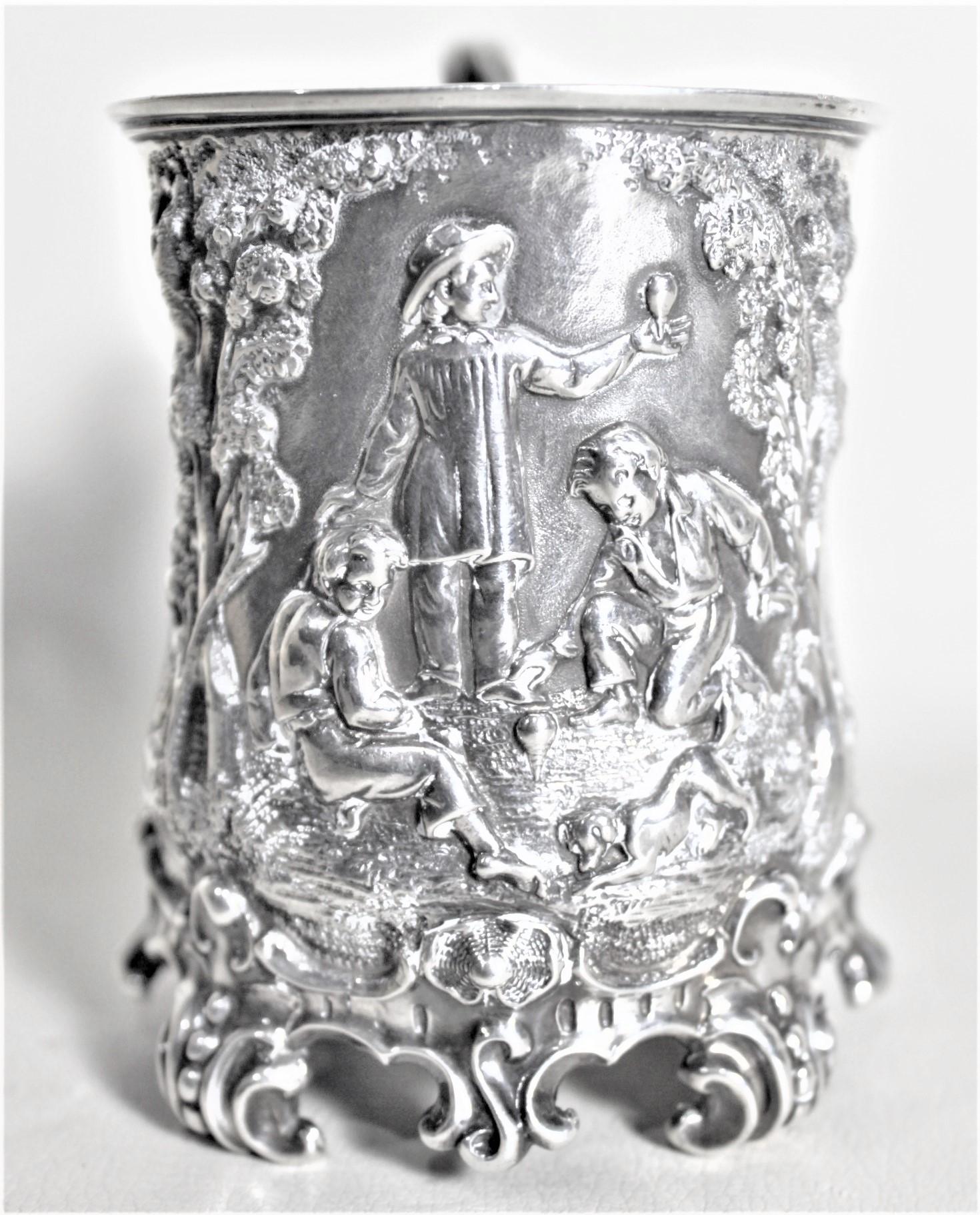 This antique sterling silver Christening cup was made in England in approximately 1848 in the period Early Victorian style. The front of this sterling cup is ornately decorated with a detailed vignette of three boys in a forest playing with spinning