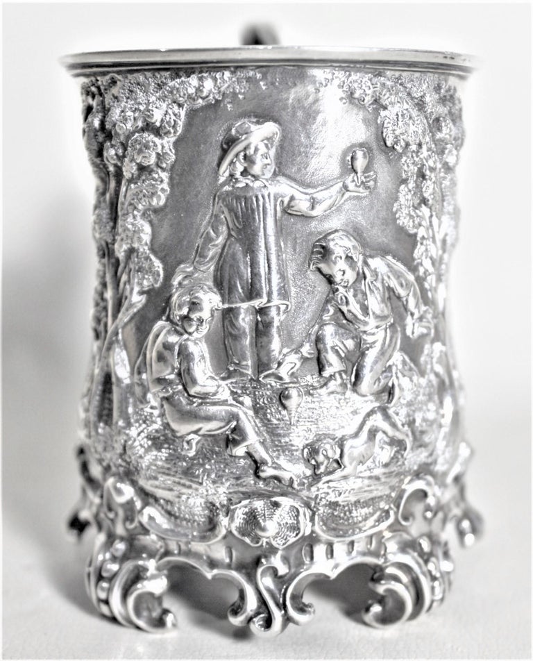 This antique sterling silver Christening cup was made in England in approximately 1848 in the period Early Victorian style. The front of this sterling cup is ornately decorated with a detailed vignette of three boys in a forest playing with spinning