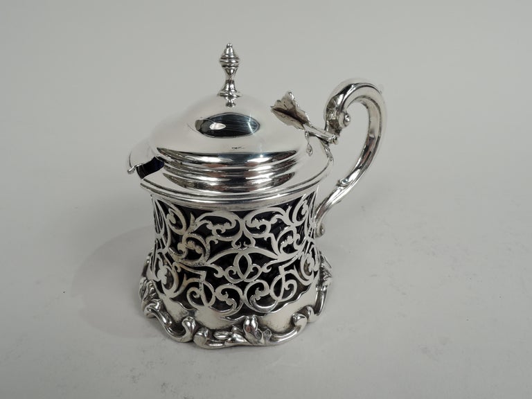 Victorian sterling silver mustard pot. Made by George Frederick Pinnell in London in 1844. Round with open scrollwork sides and spread base with applied leafing scrollwork. Hinged and domed cover with vasiform finial leaf thumb rest. Capped s-scroll