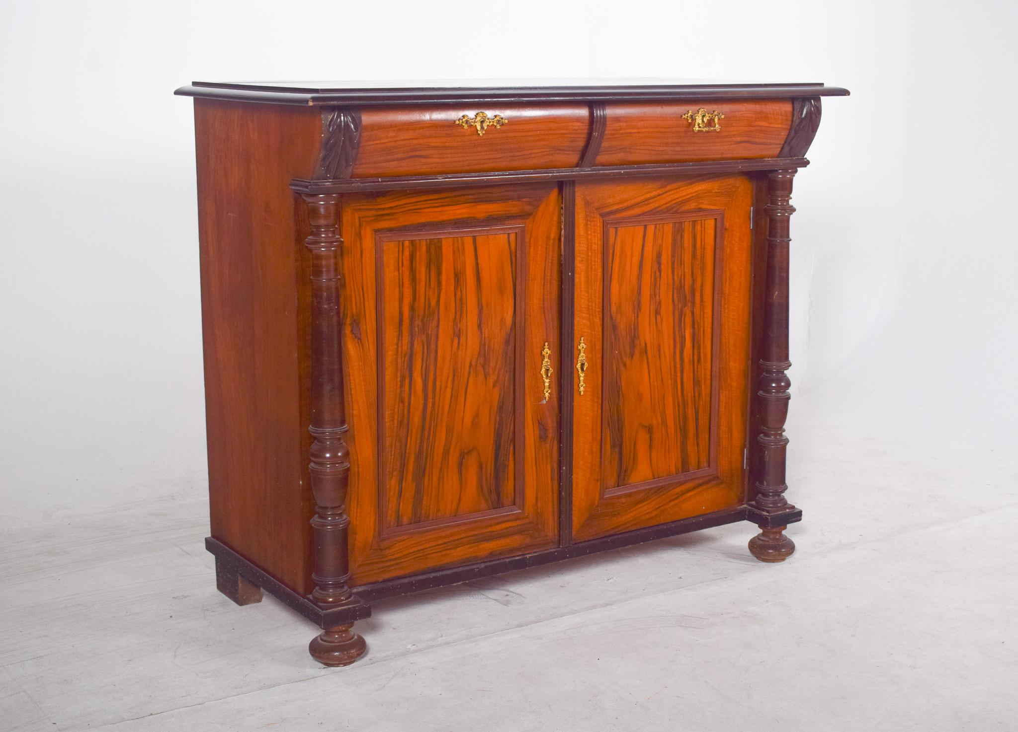 Tall sideboard in walnut. With classic lines and a sturdy structure, resting on turned feet in the shape of a flattened ball, it has two upper drawers with worked gold metal handles and two lower doors with a recessed panel, revealing the beautiful