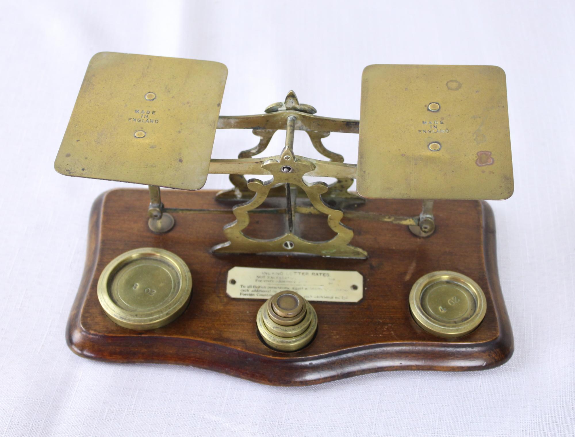 A small English postal scale, identified as circa 1915 given the prices per oz. and the foreign country assignations on the plaque. It stands on an oak base with serpentine edge. There are 6 weights which measure 8 oz, 4 oz, 2 oz 1 oz, ½ oz, ¼ oz.