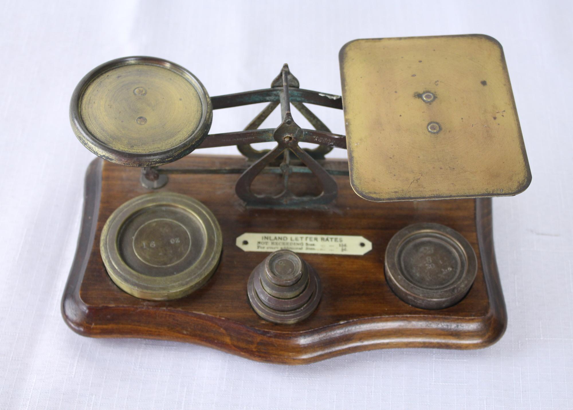 A small antique brass and wood postal scale. It stands on an oak base with serpentine edge with attractive bun feet. There are 6 weights which measure 16 oz, 8 oz, 4 oz 2 oz, 1 oz, 1/2 oz. Note: We could not find a maker’s mark on the piece, but it