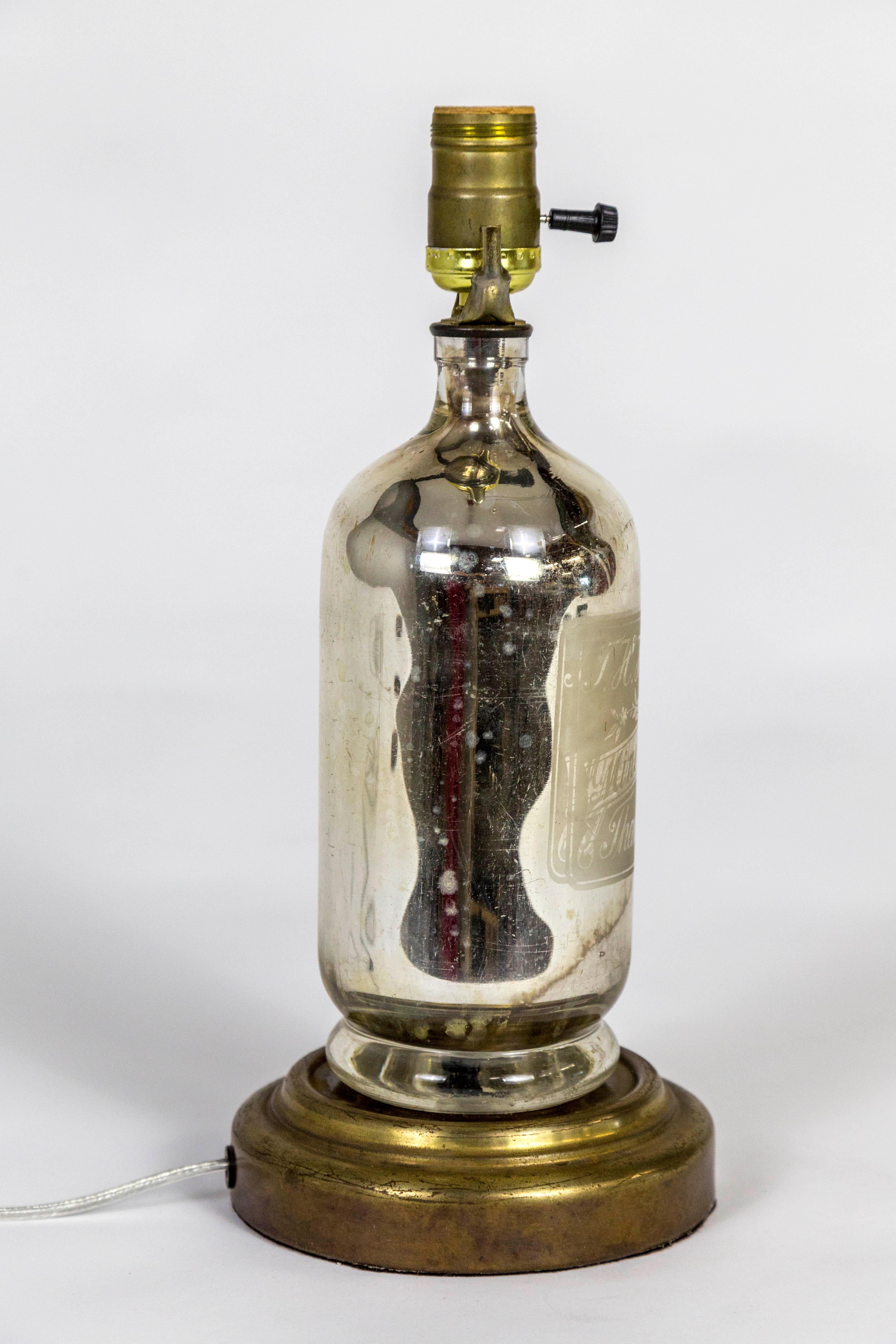 An antique, mercury glass bottle made into a table lamp with a perfectly paired, aged, gilt base. The bottle is etched with a label for mineral water by J. H. Hawkes, Thames Ditton. Newly wired with an inline switch. 13.5