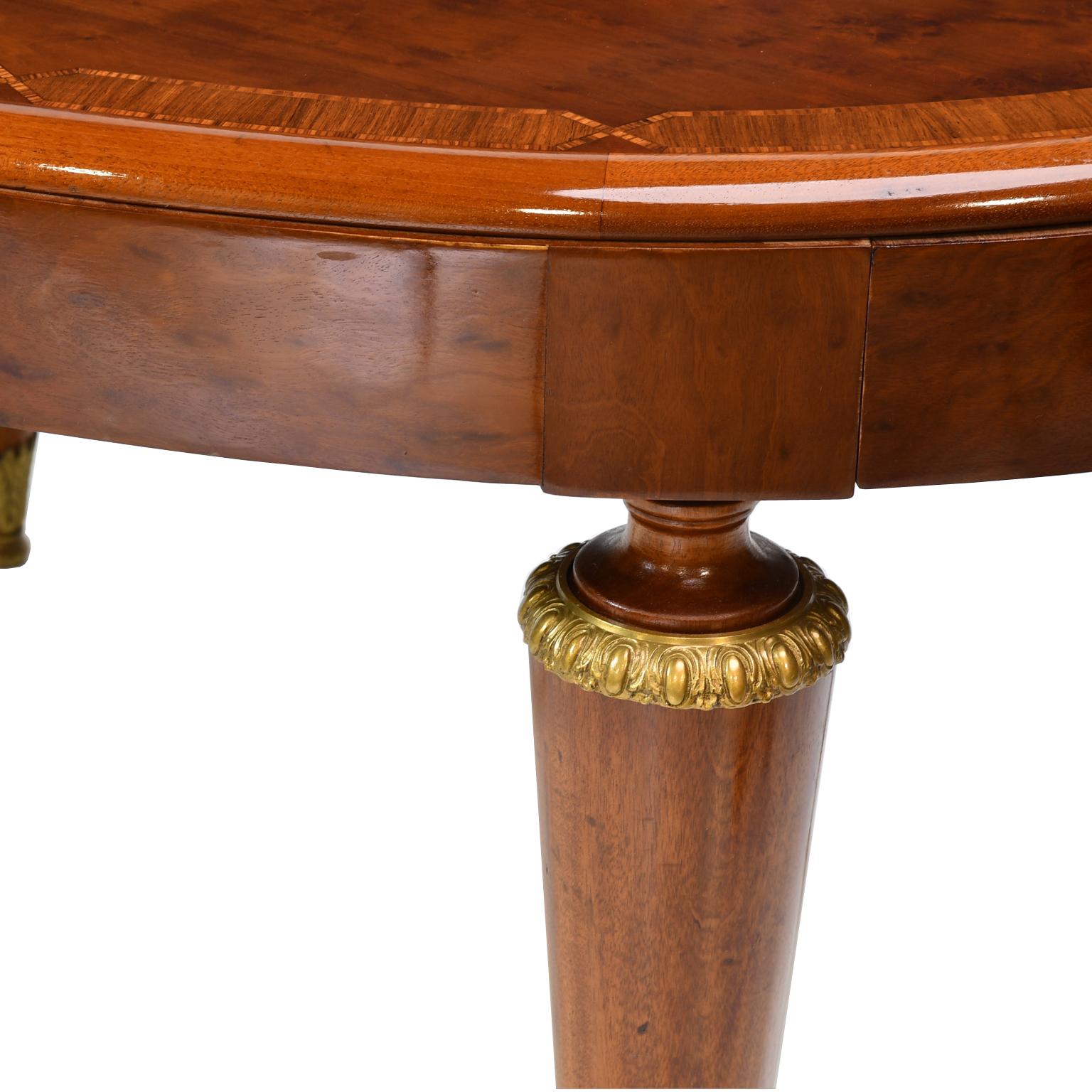 A sleek and stylish French Art Deco dining table in polished quilted plum mahogany with inlays of kingwood and satinwood along the banded edge. This fine table has an oval top with an apron over a base with four inverted conical legs with bronze