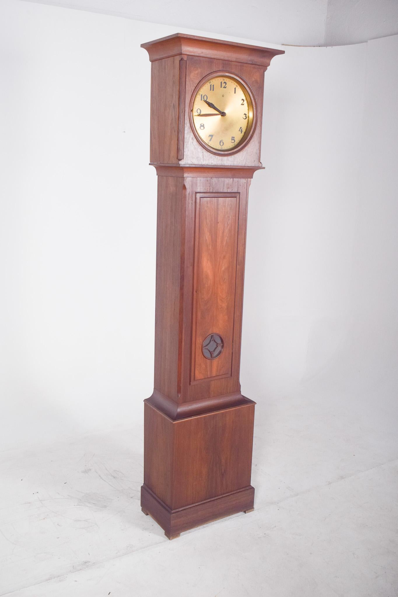 Grandfather clock. In varnished wood, with a smooth finish and warm tone. The circular gold dial features black Arabic numerals and simple hands, with a classic and elegant design. The top of the cabinet, where the dial is located, has a slightly