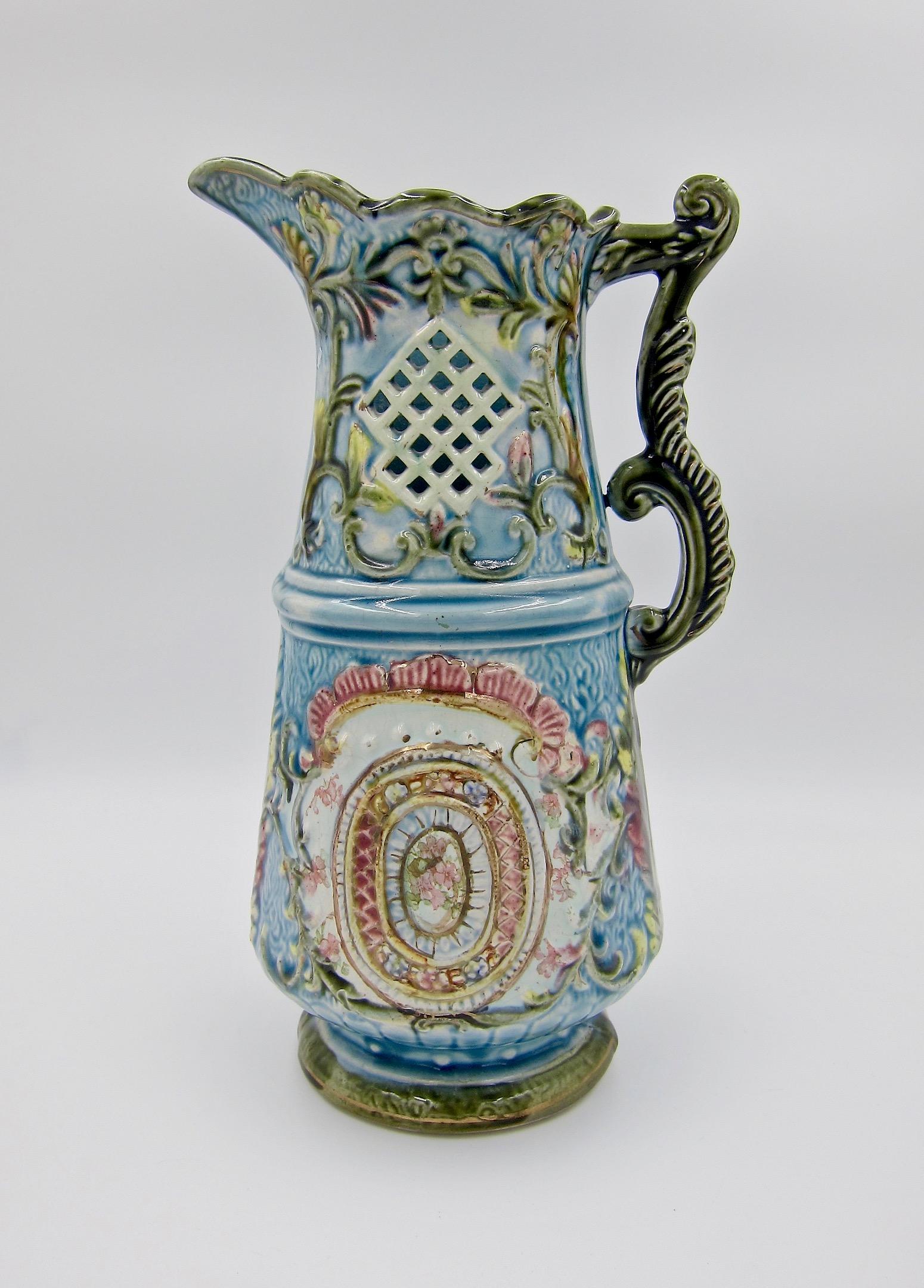 An ornate late 19th century European Majolica pitcher on a spreading foot with scrolling handles and pierced, diamond pattern accents. The Victorian era design includes molded flowers, foliage and shell motifs hand-painted in pink, yellow, and green