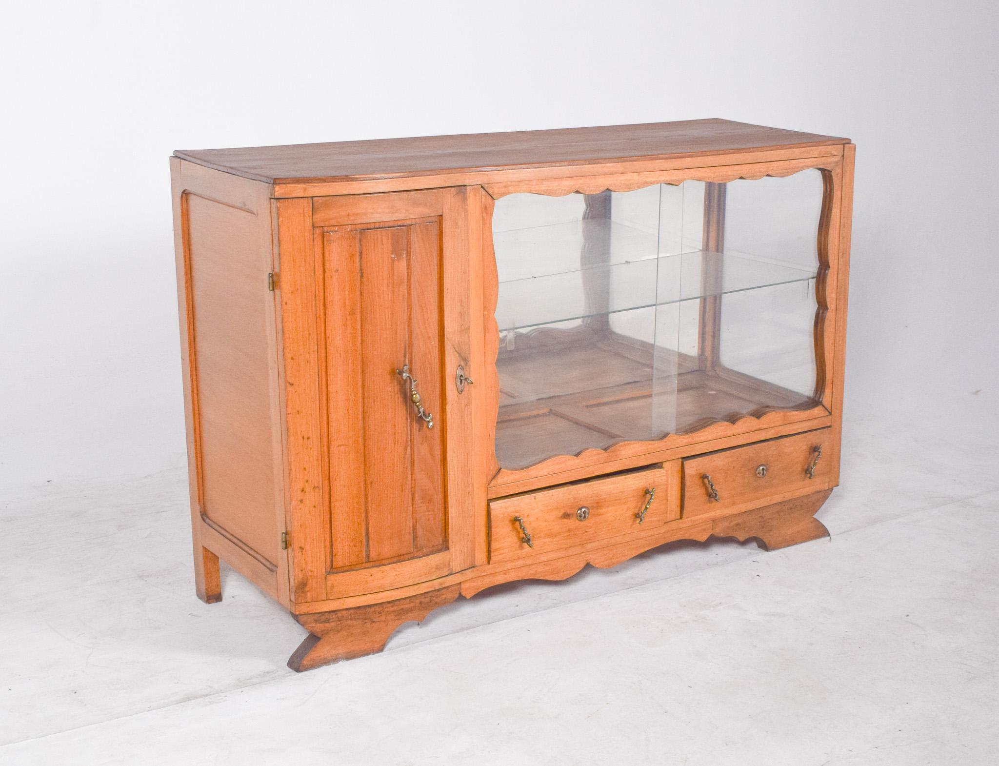 Sideboard with display case with one door and two drawers. With an asymmetrical body, it has a wooden door on the left and a large glass display case on the right, framed by undulating contours that soften the look. Inside, two glass shelves create