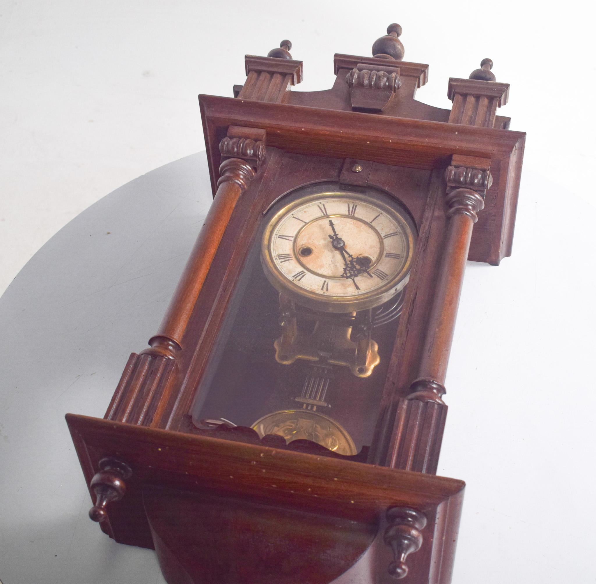 Reloj de pared antiguo. La pieza es de madera oscura, con detalles tallados y columnas laterales torneadas. La parte superior presenta elementos decorativos simétricos, como pequeños pilares y un ornamento central redondeado.
La esfera del reloj