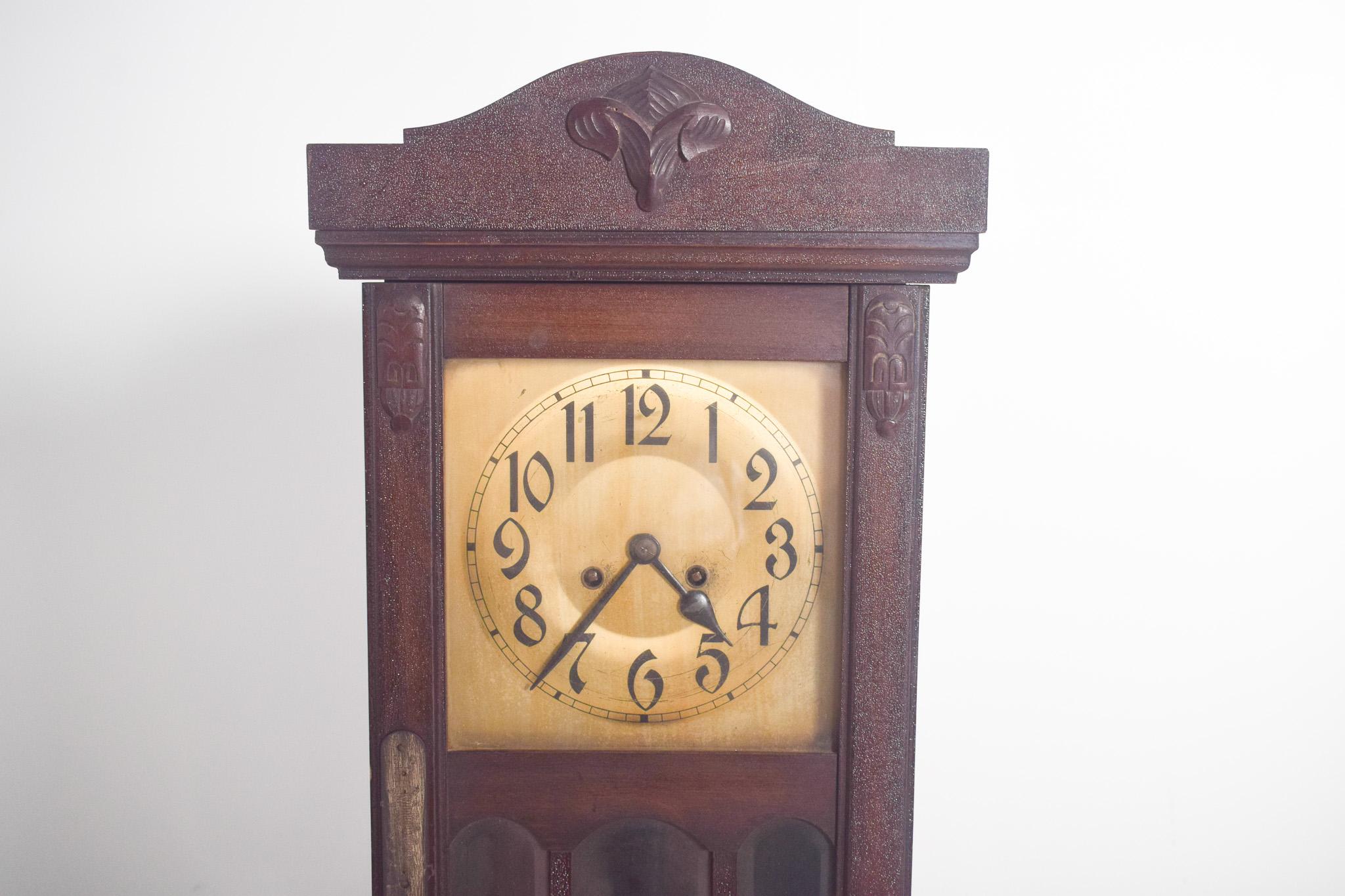 Horloge murale, ancienne. Dotée d'un design classique et sculpté, d'une structure rectangulaire et robuste, elle présente un cadran à fond clair, des chiffres arabes noirs et de belles aiguilles. La vitre frontale protège le mécanisme interne et