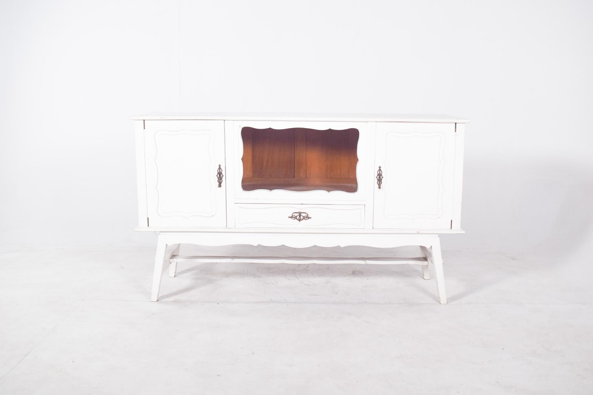 Sideboard painted white. The sideboard is marked by sinuous lines in the door cut-outs and the central niche, as well as the sloping feet that support a base with a connecting bar. The machined metal centre handle completes the composition with a