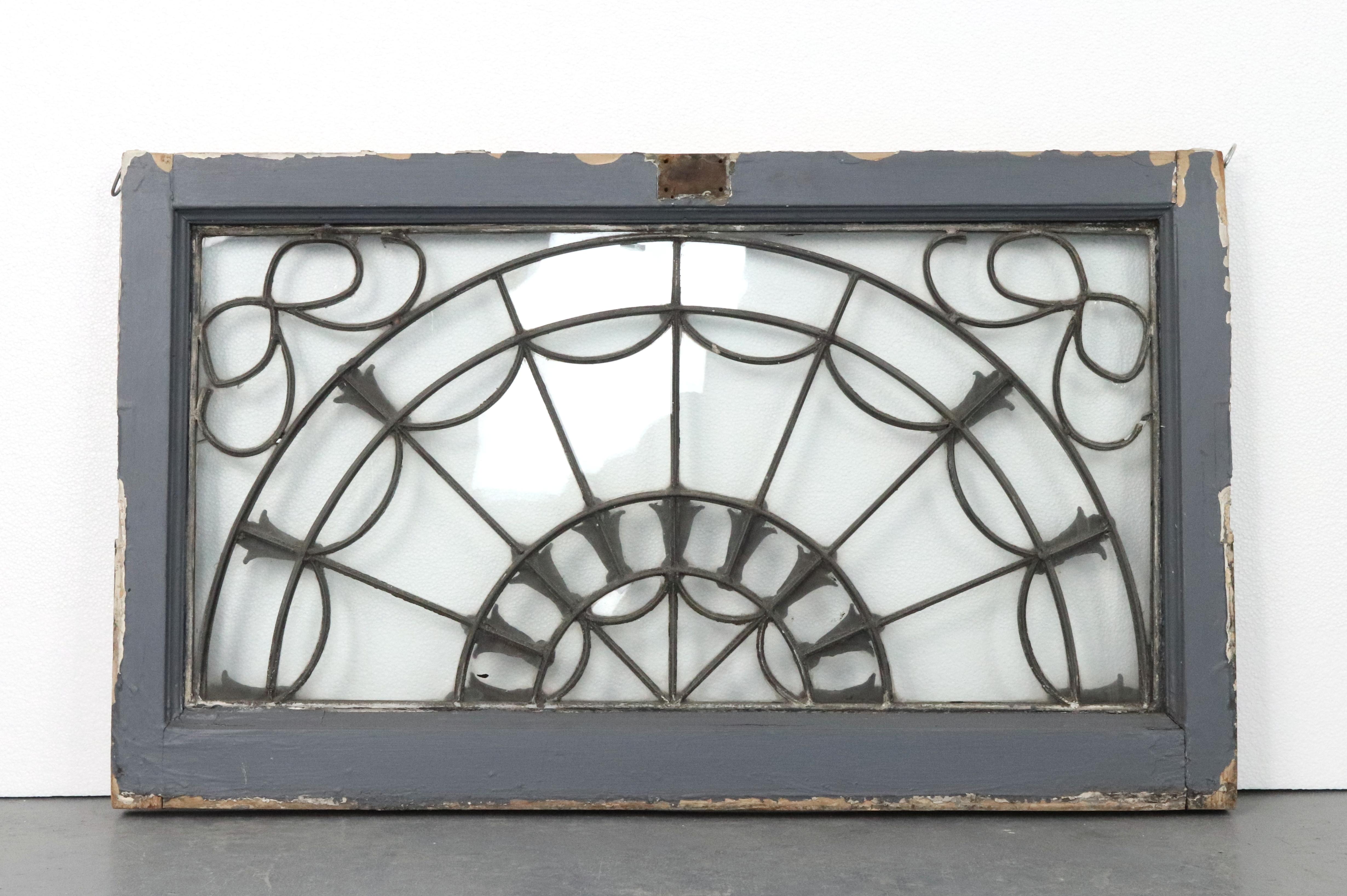 Antique wood frame fanlight transom window with a fan like design with raised floral elements at the intersections. The frame is painted black on one side and gray on the other. Fanlights are a specific type of transom window with a semi-circular