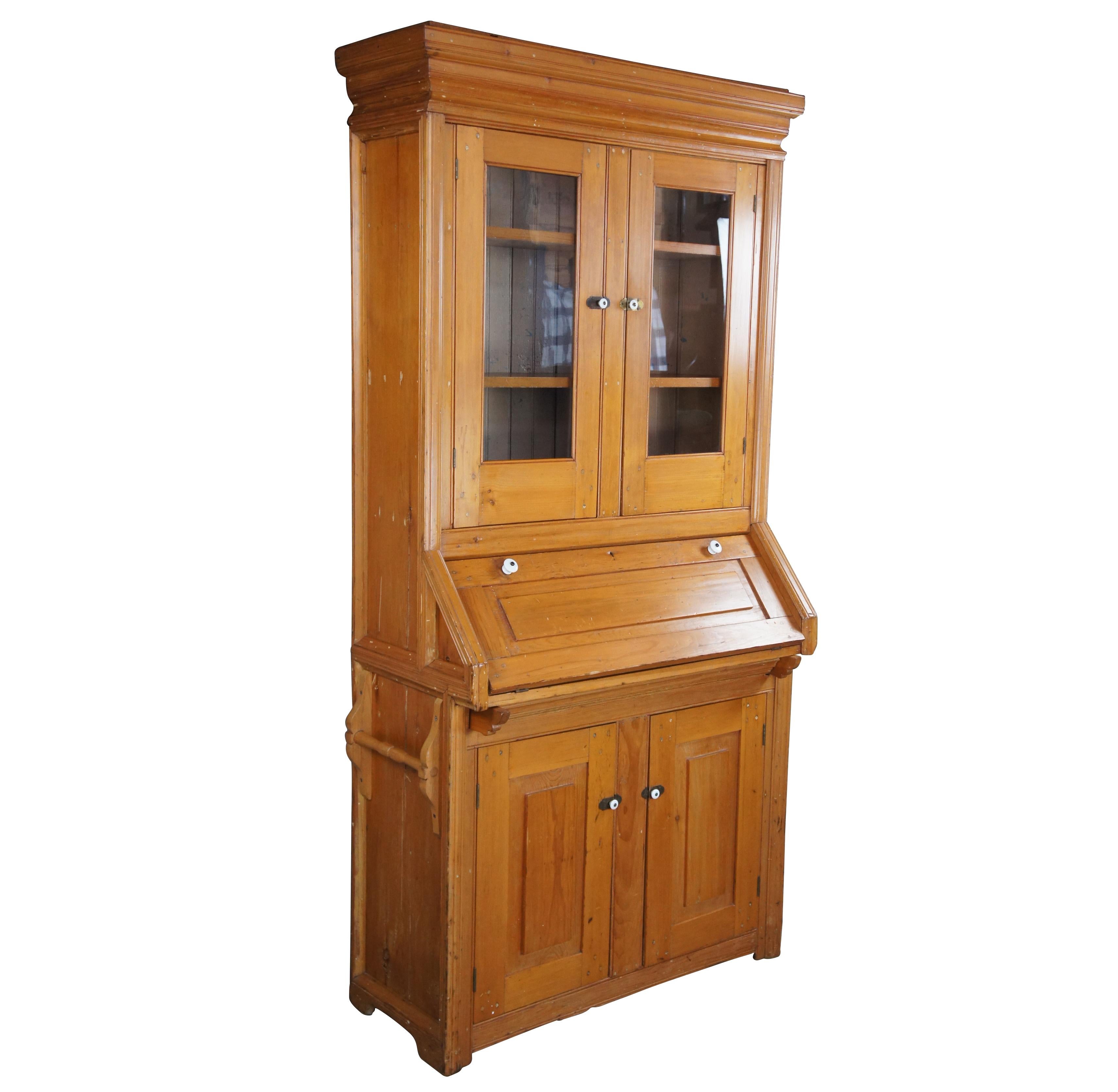 An early 20th century pine secretary desk or cupboard. Features an glass covered upper bookcase with two shelves, drop front writing surface with shelf and lower cabinet with shelf for accessory storage Cabinet and door pulls are made from brass and