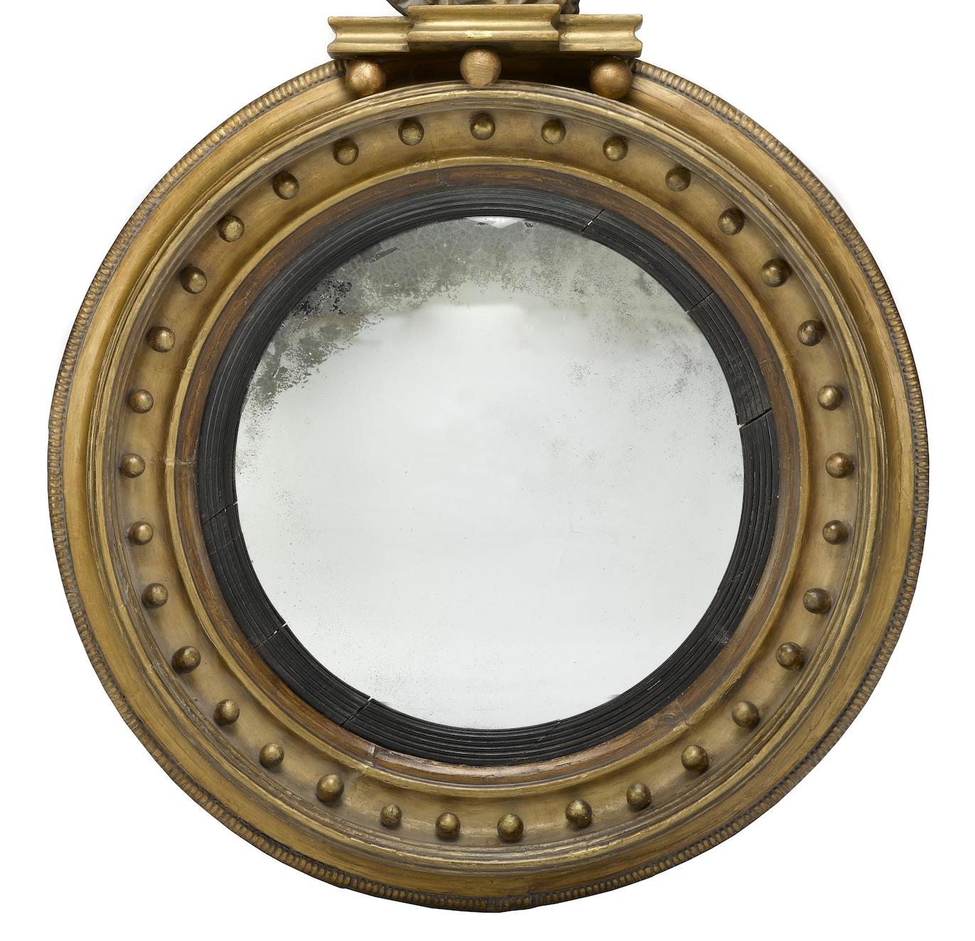Presented is a Federal period bullseye mirror with a stunning eagle embellishment at top. The mirror frame is hand carved, constructed of a hardwood, and overlaid by gold leaf. At top is an eagle perched on a small Doric column. The mirror frame