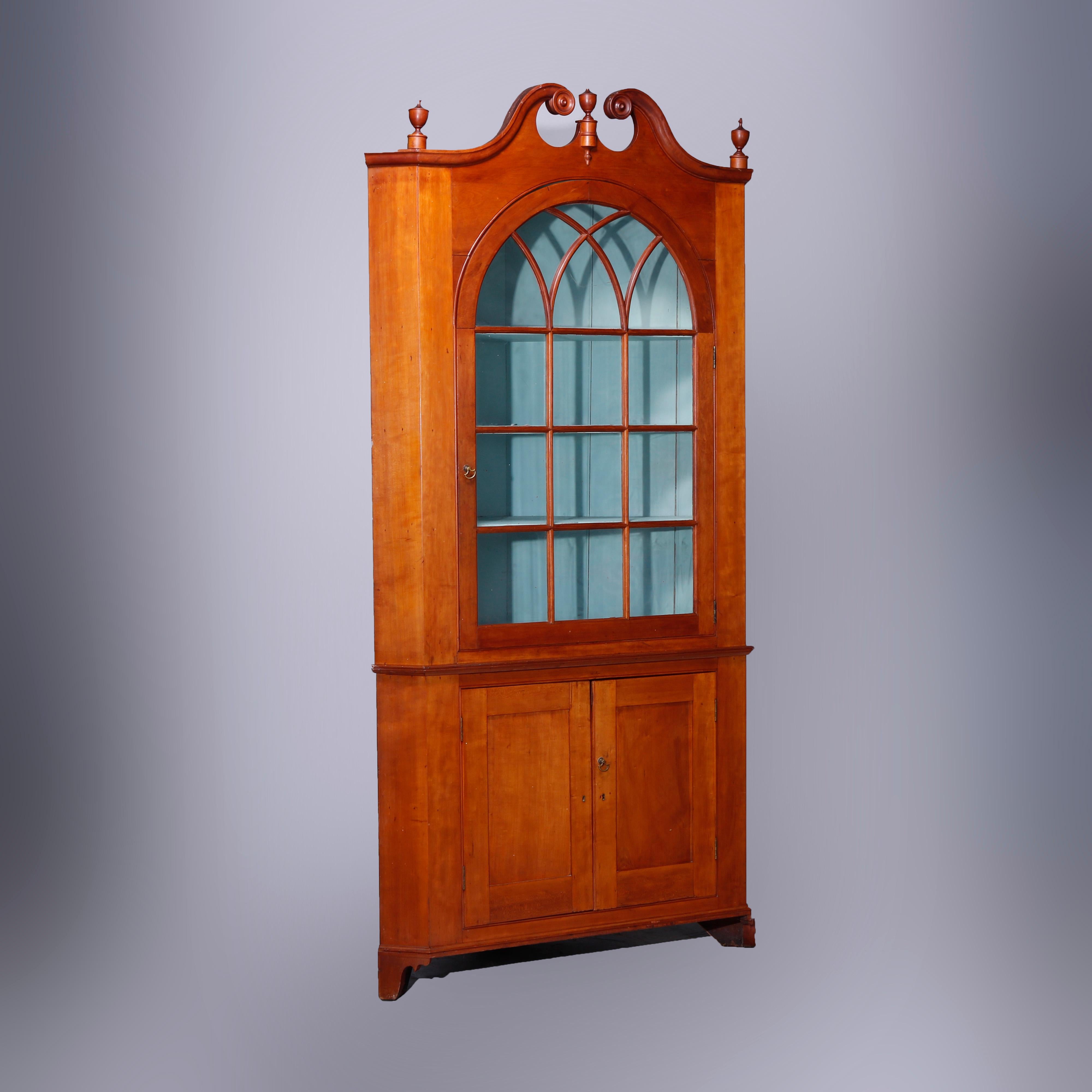 An antique Pennsylvania Federal two-piece corner cupboard offers cherry construction with broken arch crest having central finial surmounting cabinet with upper having single door with arched mullioned glass window and opening to shelved interior