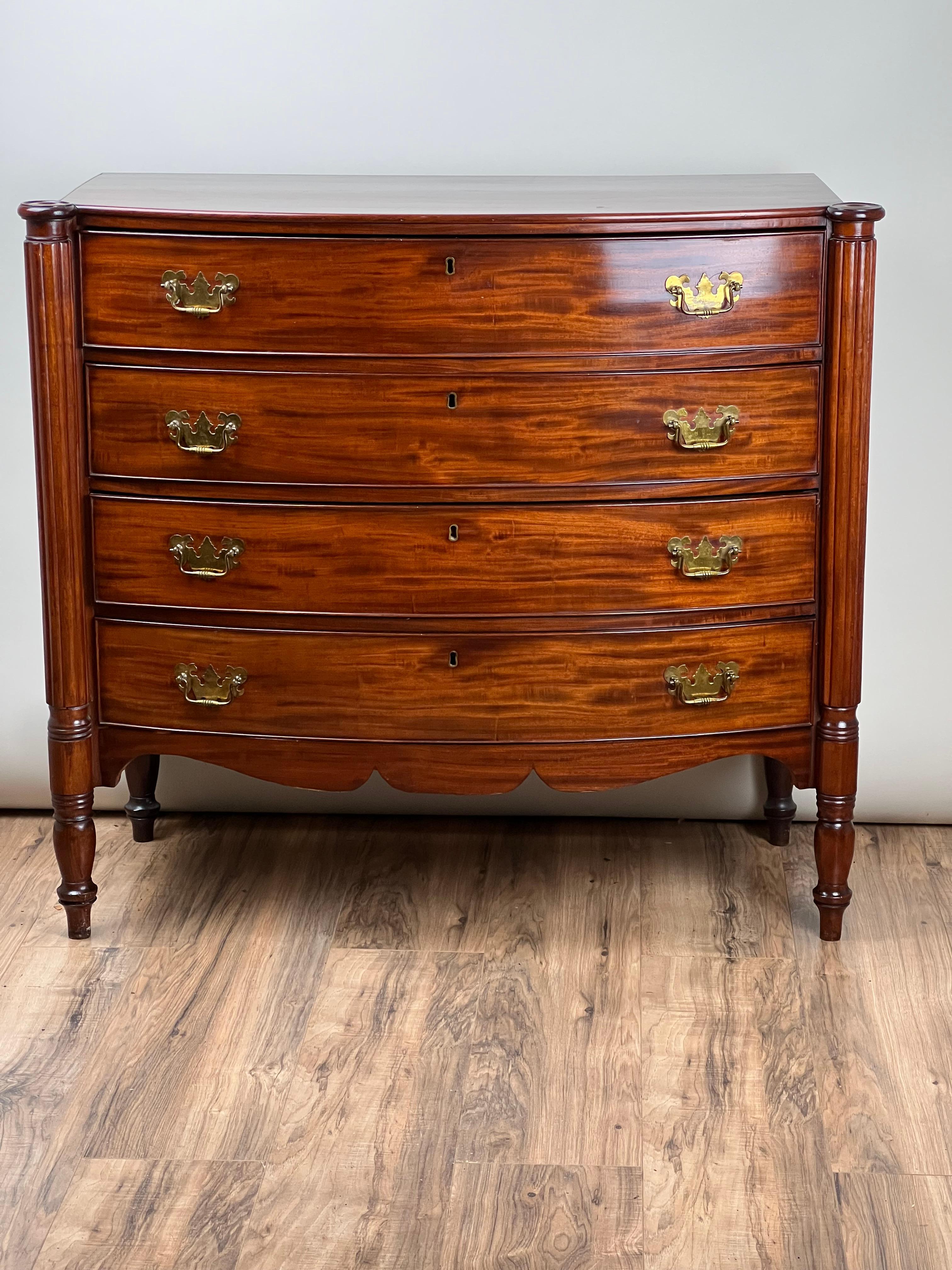 A classic of early 19th-century American craftsmanship, this Federal period Sheraton chest was made around 1830 and showcases richly figured solid mahogany. With its gracefully bowed front, turned legs, and reeded side columns, the chest offers a