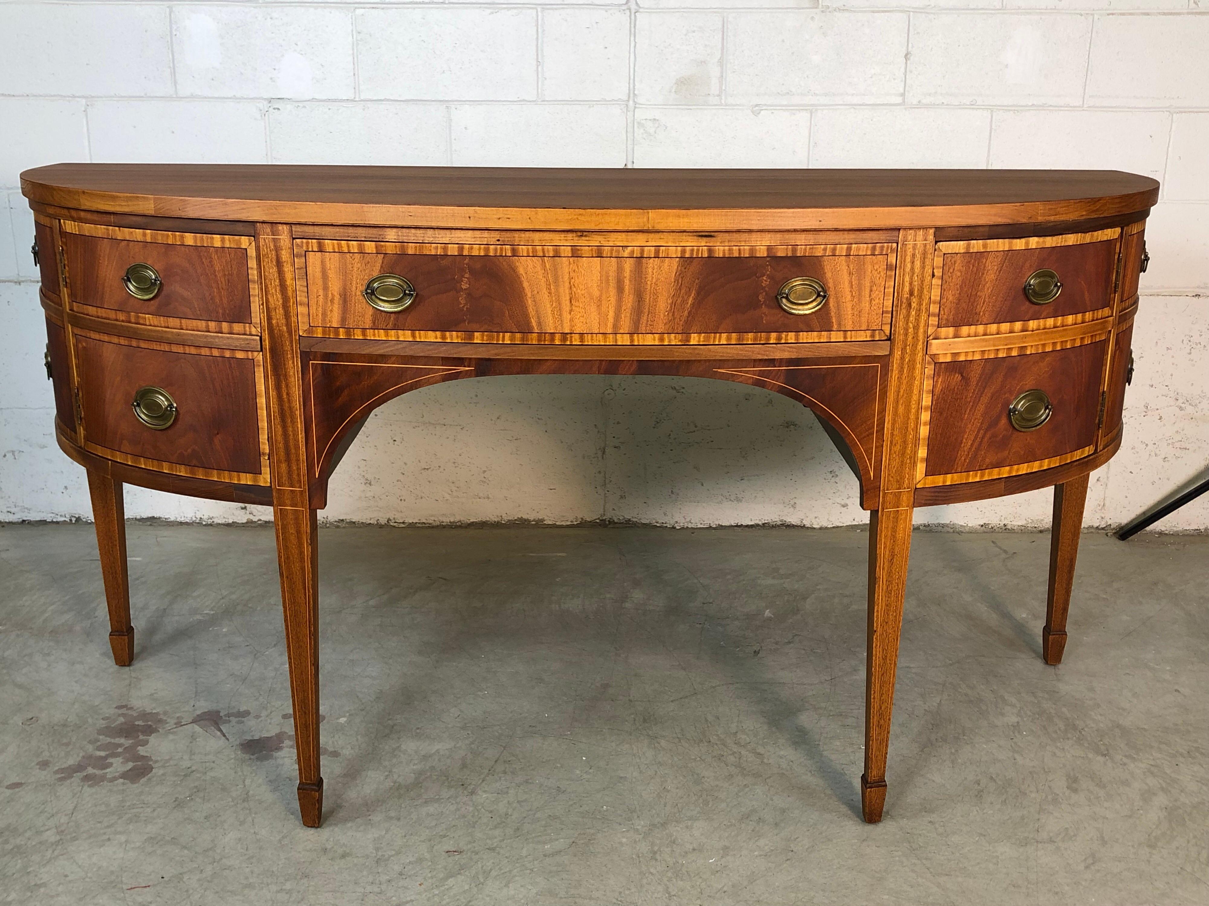 Antique Federal style inlaid mahogany demilune sideboard. The sideboard has matchbook style mahogany fronts with brass pulls. There is one center drawer for storage and another draw hidden behind a side door. The side drawers are faux and there is