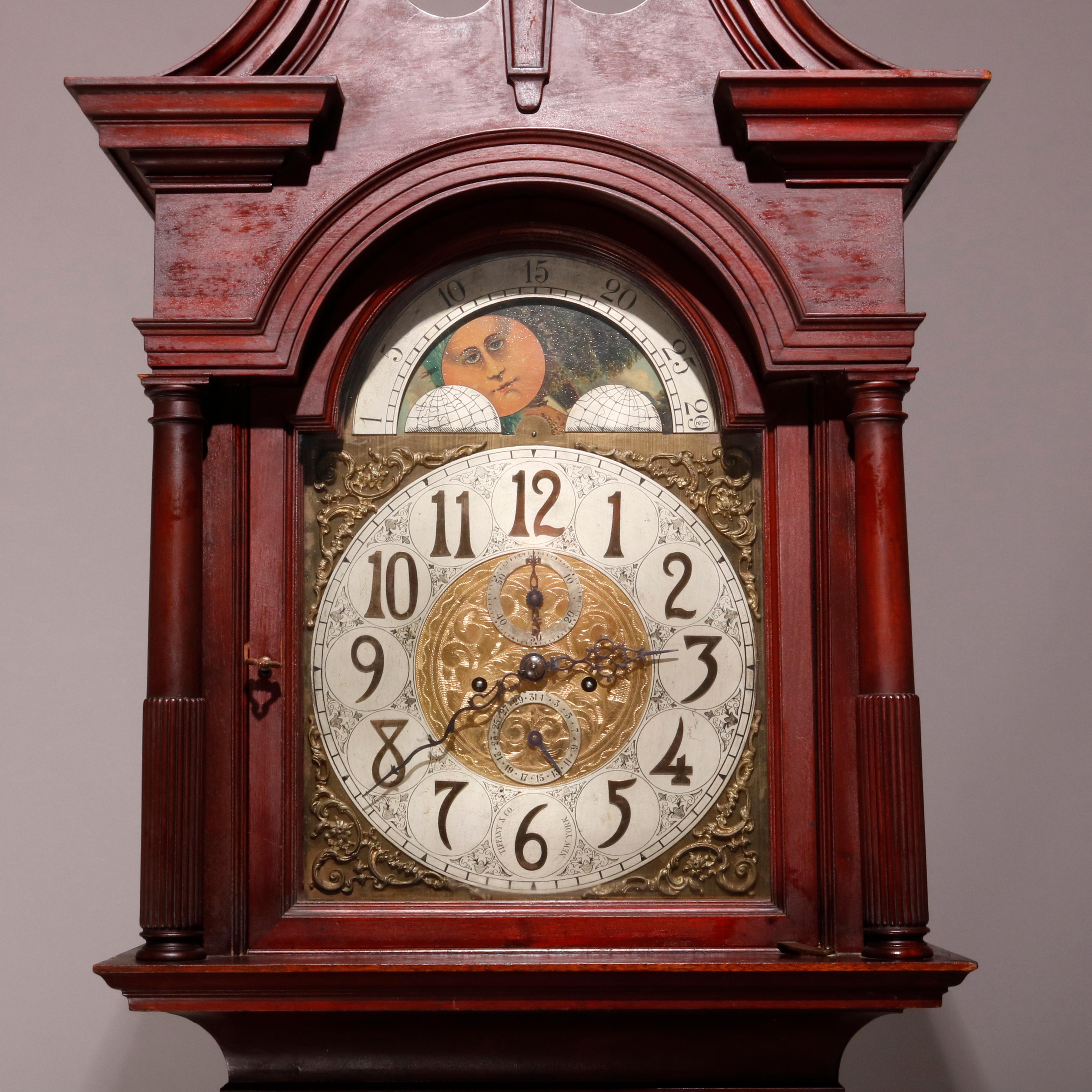 An antique Federal style long case clock by Tiffany & Co offers mahogany case with broken arch pediment having central urn form finial surmounting moon phase face with Arabic numerals, seconds and date dials, flanked by Doric columns with reeded