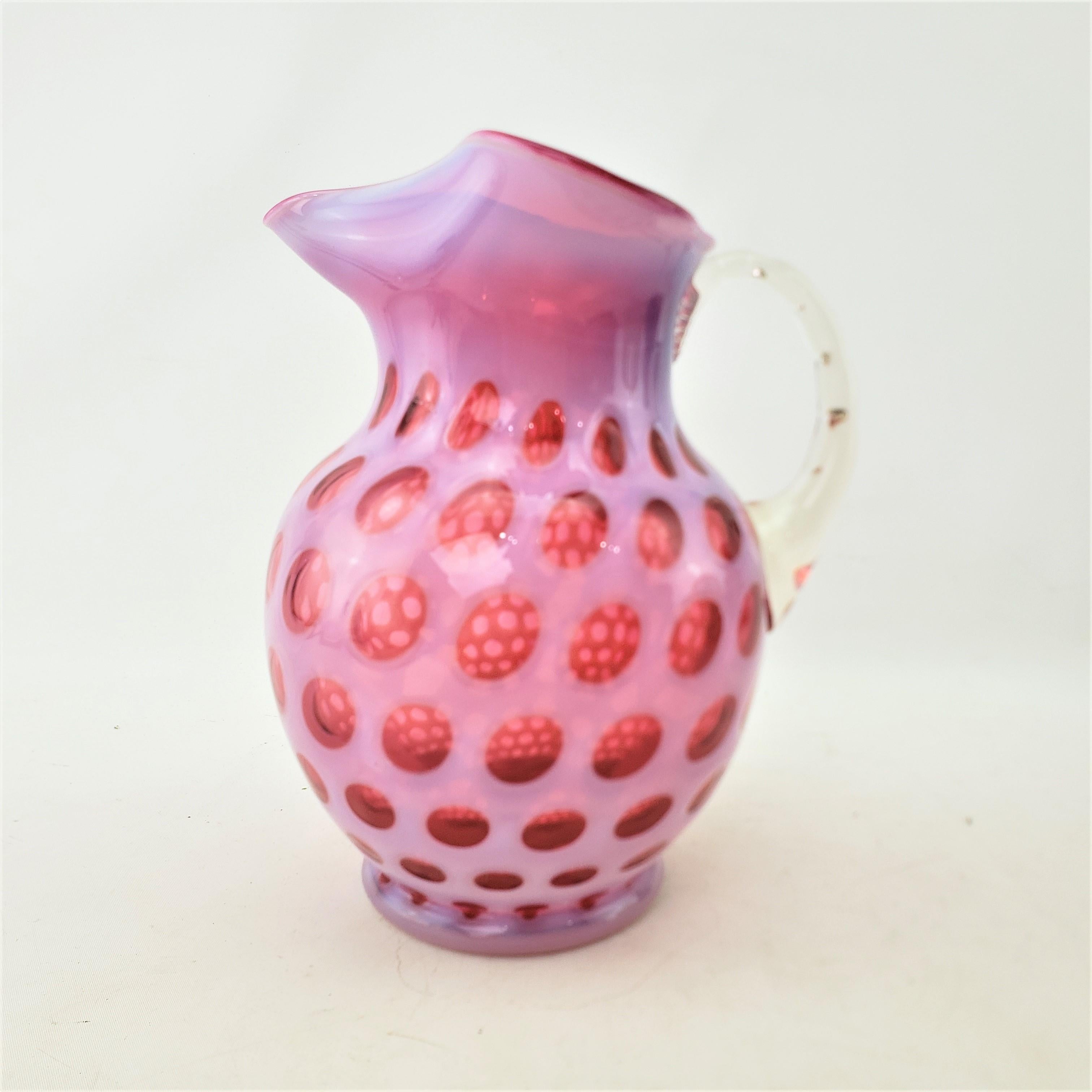 This antique pitcher is unsigned, but presumed to have been made by the Fenton Glass Company of the United States and date to approximately 1900 and done in a Victorian style. The pitcher is composed with a hand-formed cranberry glass base with an