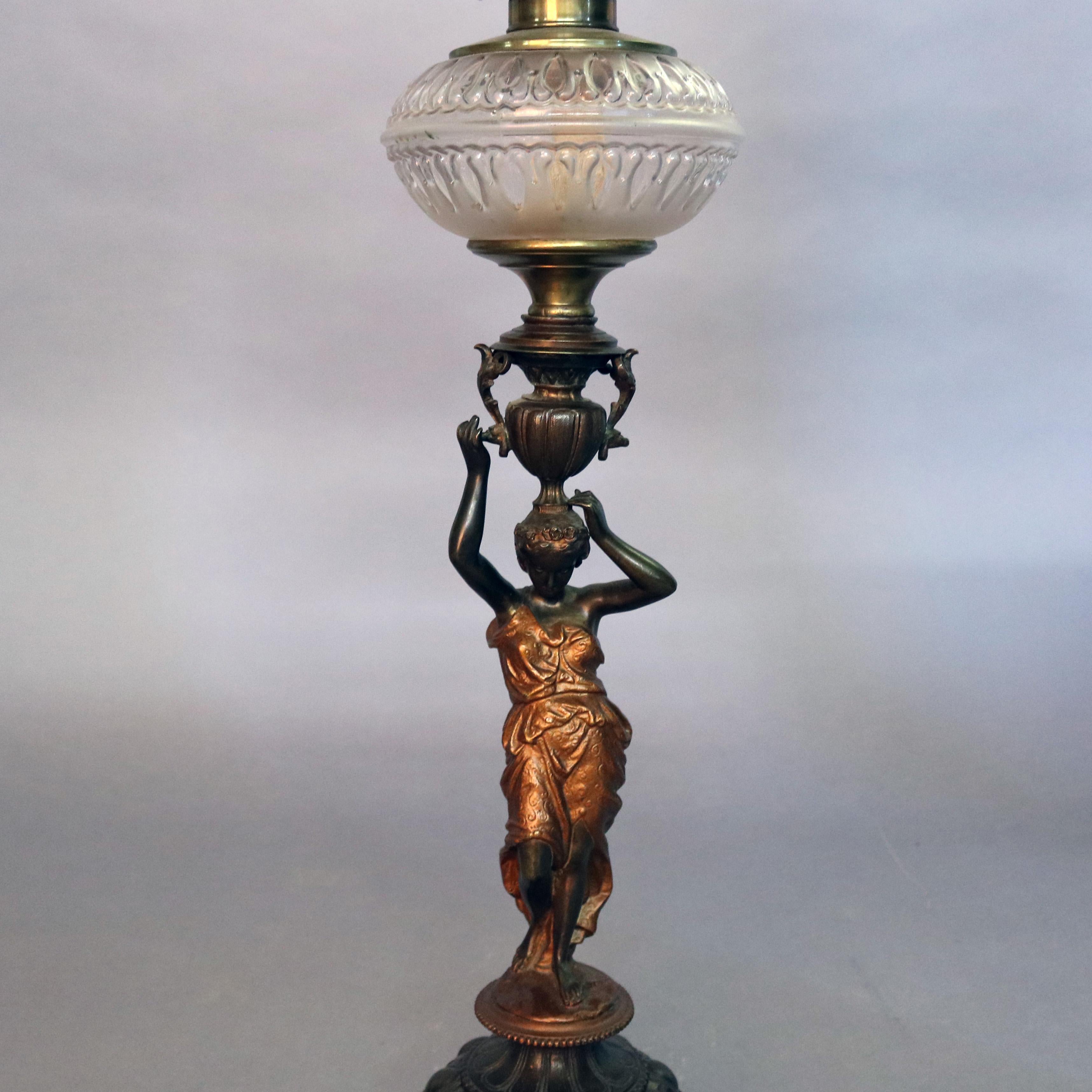 An antique gone with the wind figural lamp features cast metal base with statue of classical woman with gilt dress and urn raised on foliate tripod base surmounted by oil lamp with pressed glass font and etched cherub decorated shade, circa