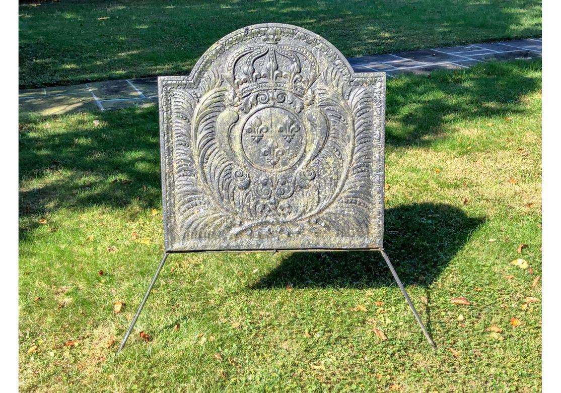 An arch top black fireback with relief decoration of palm fronds framing a crown and crest with fleur-de-lis motifs. With a leafy outer border. Incrustations overall. Custom iron stand is included. 

Measures: 31 x 32