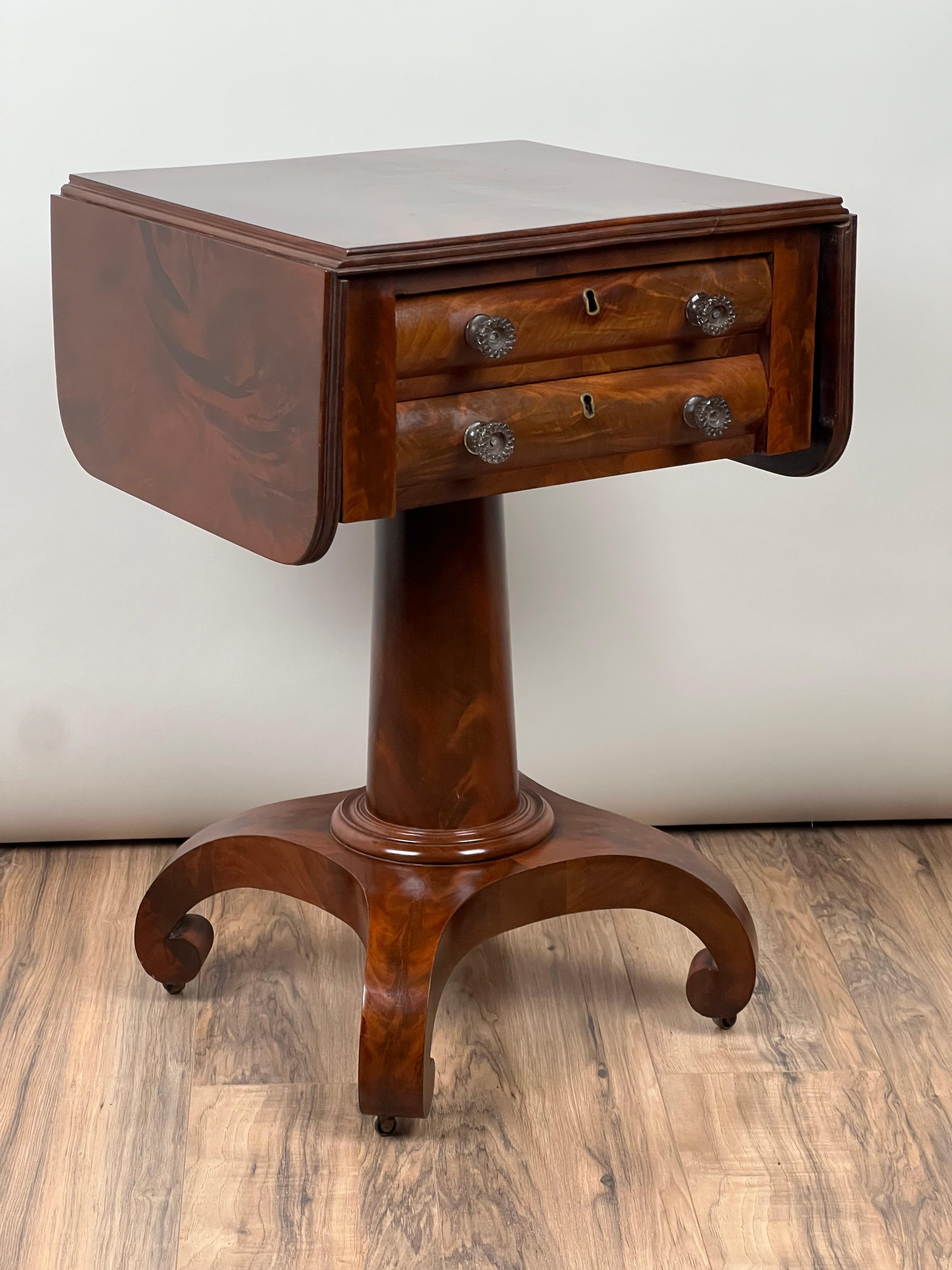 This striking American Empire two-drawer work table, dating to around 1840, features a beautifully figured flame mahogany veneer that lends a rich depth to the piece. The clean, straight lines of the table’s form are elegantly complemented by the