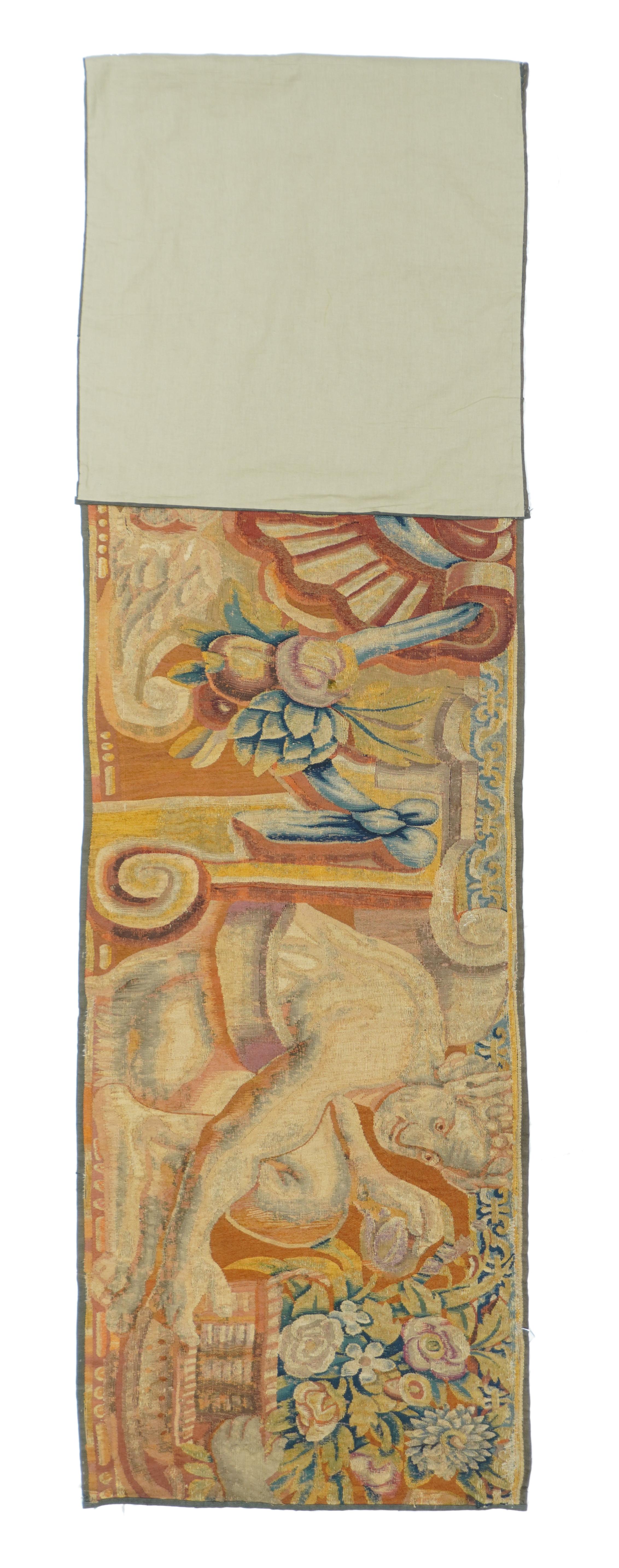 From a Brussels historical or mythological tapestry, matching our 229.
Two crouching male figures hold overflowing flower baskets, and flank a shell-like central motif with attendant fruits and a winged cherubim head below. Trompe l’oeil volute