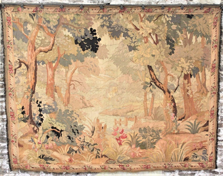 This large tapestry is unsigned but believed to have been made in Belgium in circa 1895 in the country Flemish style. This tapestry is done in muted tone or verdure and depicts a lush forest framed with large trees on either side and greenery in the
