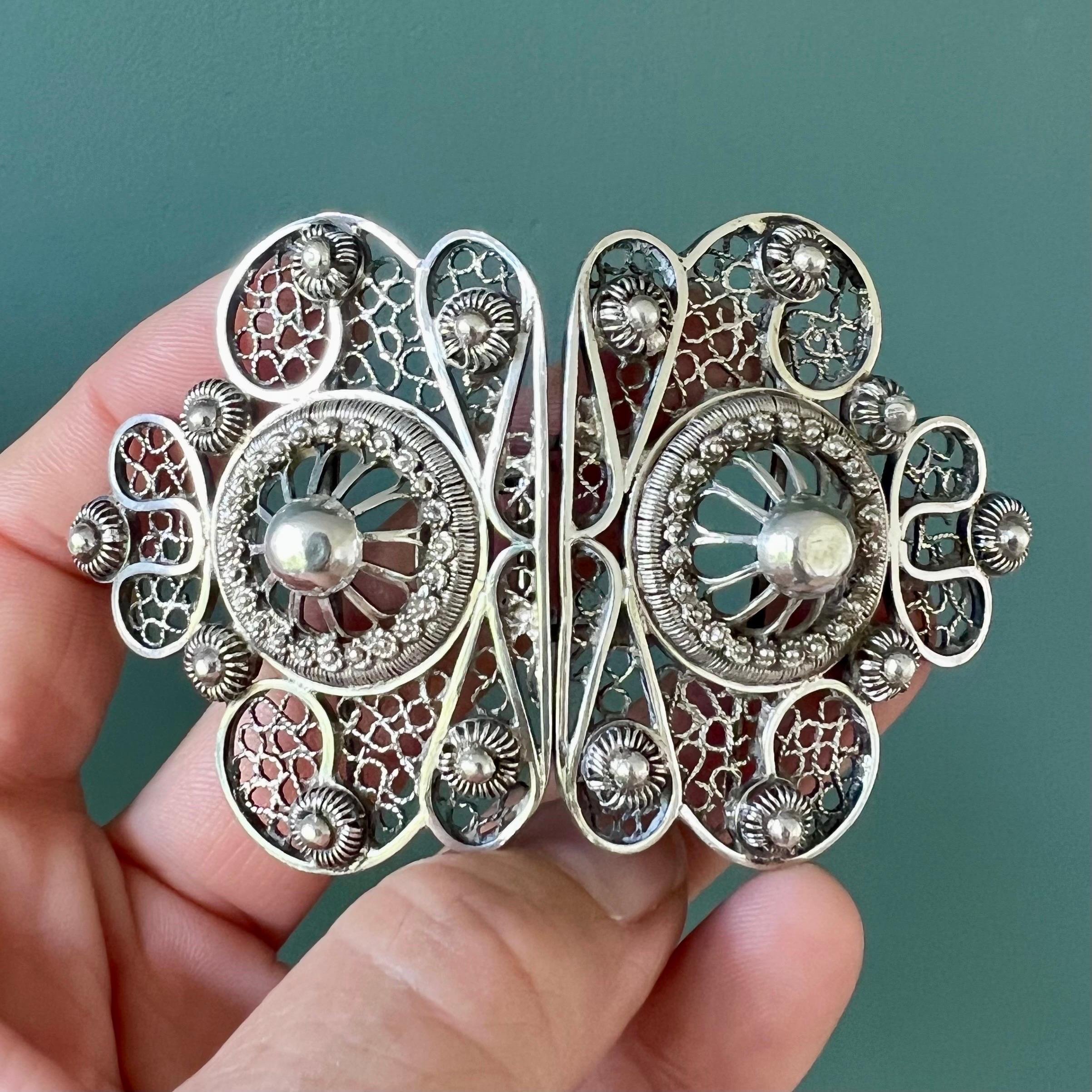 Questa fibbia da cintura in argento antico con filigrana floreale è un bellissimo esempio di intricata lavorazione artigianale. La fibbia presenta un design sorprendente incentrato su due importanti sfere d'argento, una per lato. Queste sfere sono