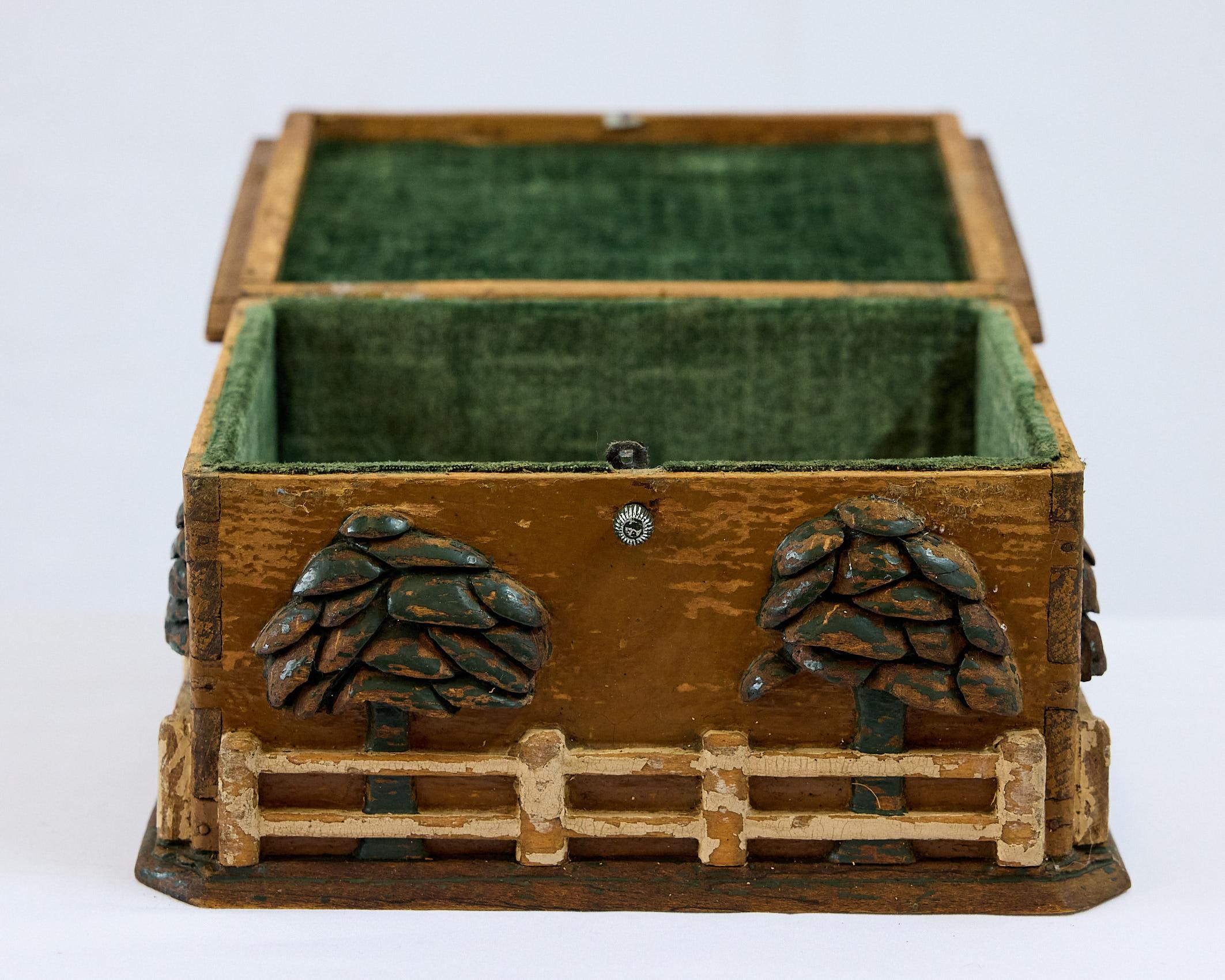 Increíble caja de Arte Popular tallada y pintada a mano con una encantadora casita con tejado de tejas en la tapa abatible. Tres de sus lados presentan escenas de árboles detrás de vallas blancas y la caja está elevada sobre una base de plataforma.