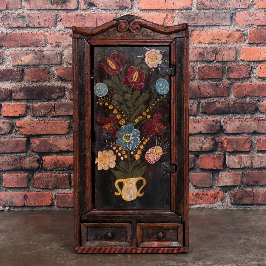 This charming tall and narrow hanging cupboard from Denmark is exceptional due to the aged patina of the Folk Art painted flowers. Take note of the carved date in the crown, the two shelves inside and the two small outer drawers. The cabinet has