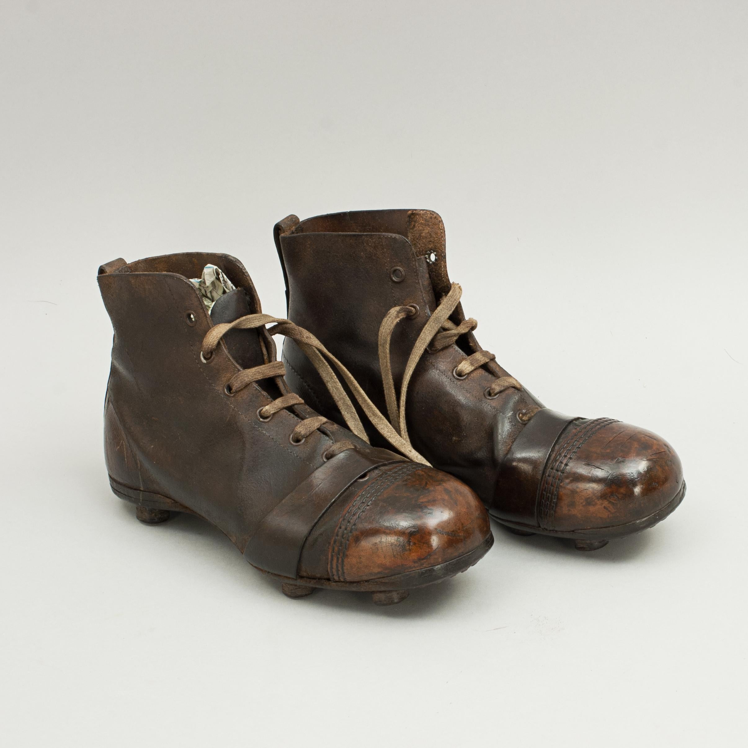 A pair of used high ankle leather football boots. The toe area on the boots are made of hardened leather with an extra leather strap behind. The sole of the boots are fitted with 6 leather studs, each stud is formed by layers of leather cylindrical