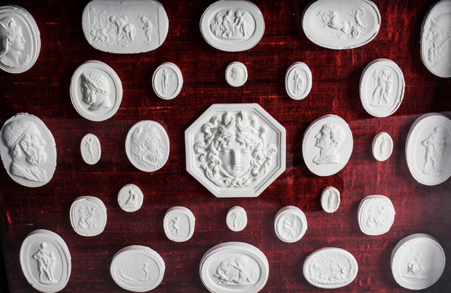 An arrangement of 29 framed plaster Grand Tour intaglios of various sizes dating from the early 20th century.

The intaglios are set in an ebonized and gilt glazed frame and mounted on a red silk ground. It consists of twenty nine plaster intaglios,