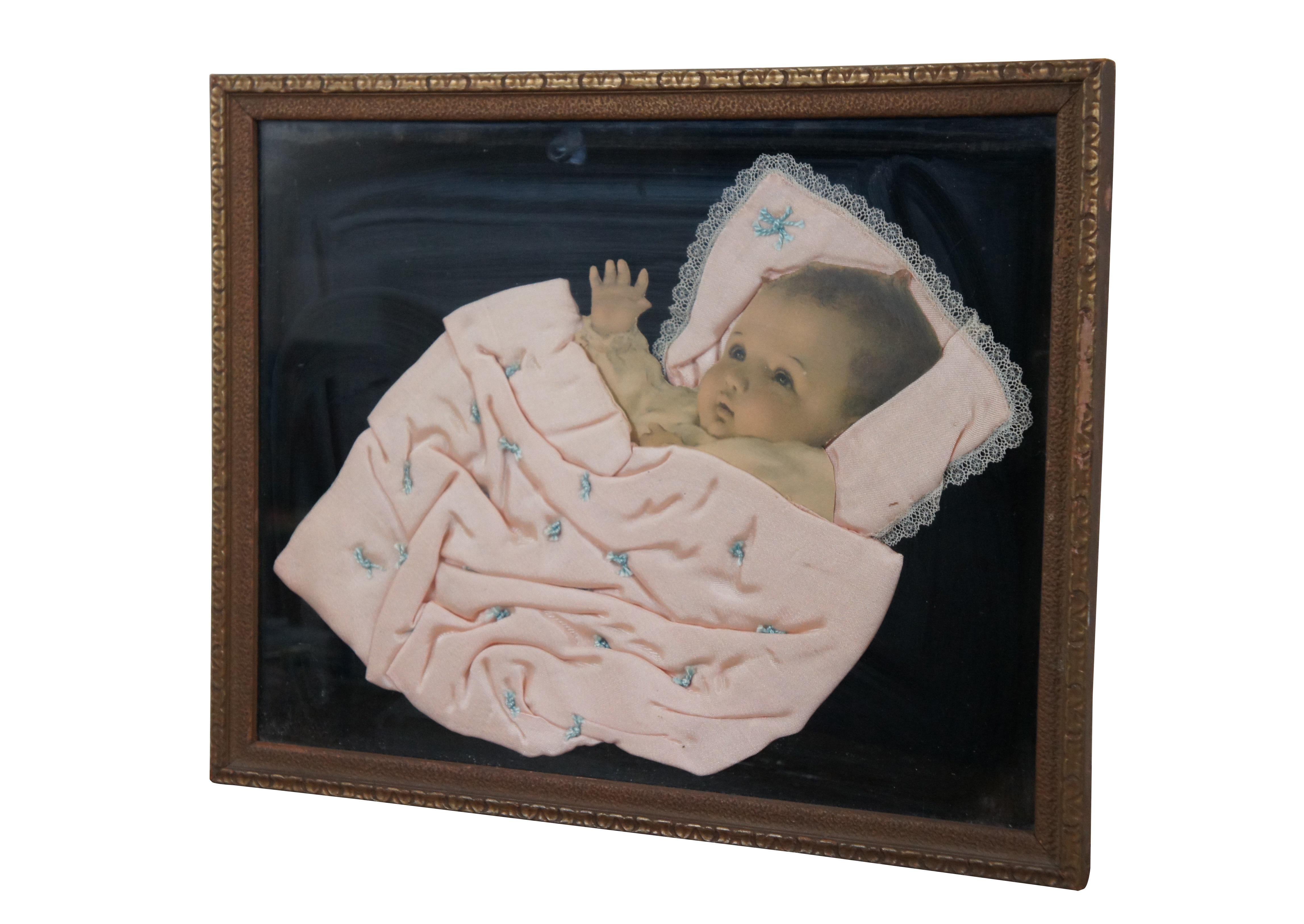 Early 20th century framed pink baby blanket with stitched blue tufting and lace trim, nestled around a die cut photograph of a baby, mounted on a backdrop of black velvet. Possibly a piece of memorial / mourning art, purportedly popular from the
