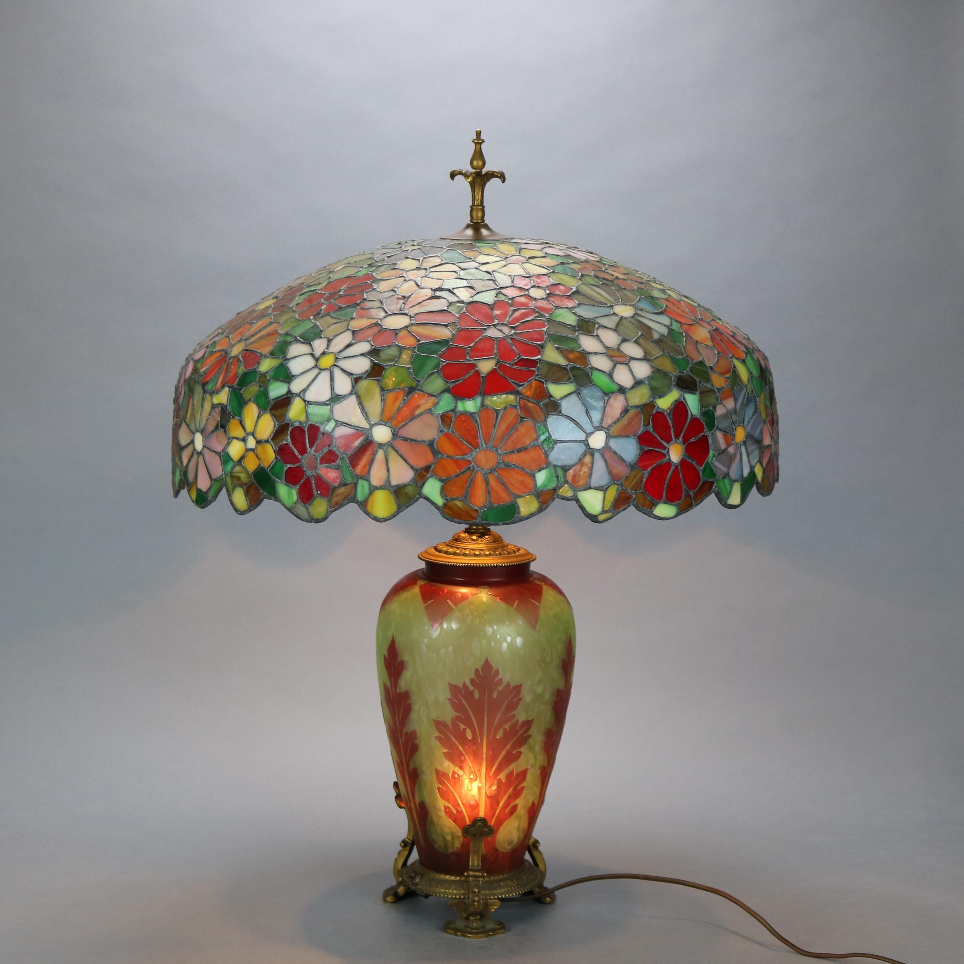 An antique table lamp offers Frederick Carder Steuben Cluthra foliate etched cutback lighted double socket base raised on gilt base with scroll feet and surmounted by floral mosaic leaded glass domed shade, c1920.

Measures: 31.5