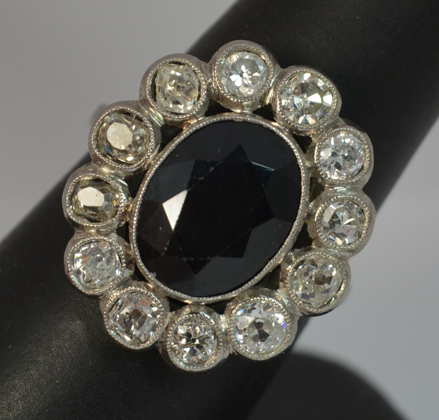 A stunning antique cluster ring.
Ring Size; R UK, 8 3/4 US
A French made example in solid platinum throughout.

Set with a large oval cut natural dark sapphire to the centre, 9.7mm x 12.5mm. 

Surrounding are twelve natural old European cut