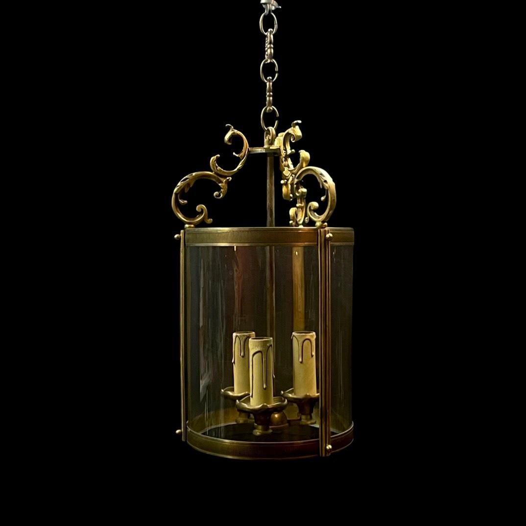 For sale is a stunning French  bronze hall lantern from the 1950s. This antique lantern features three light fittings adorned with circular bobeche drip pans, elegantly encased by sectional circular singular glass panel. It is gracefully suspended