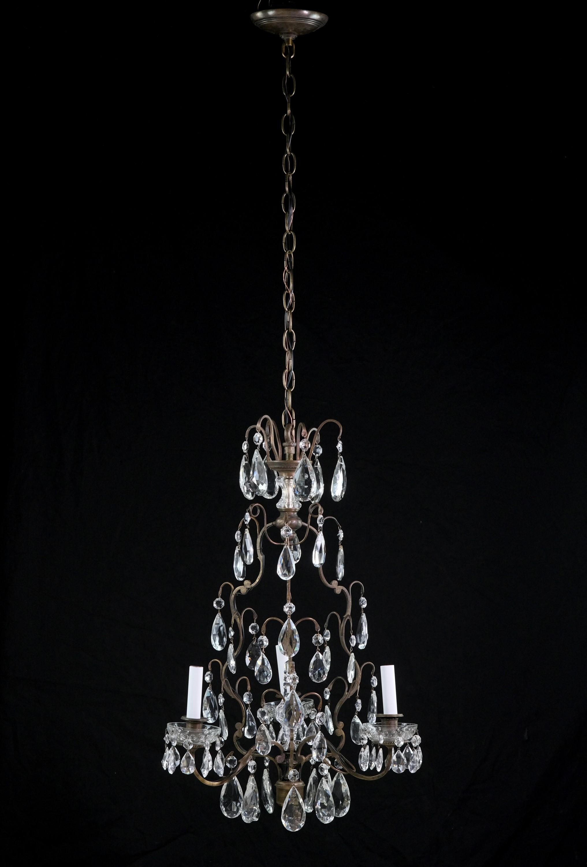This elegant early 20th century French chandelier has an elaborate bronze frame with three arms each holding a candlestick light which is adorned with a crystal bobeche and tear drop crystals. This multi-tier crystal chandelier also features a