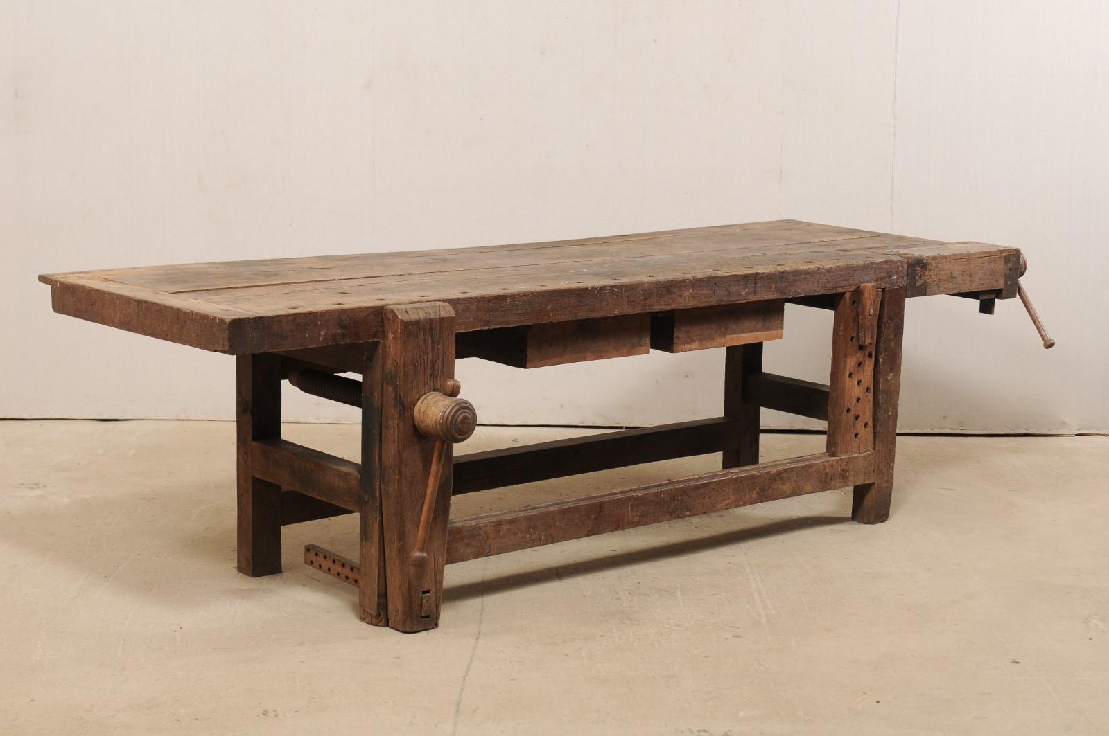 A French 9.5+ ft long work bench table with two drawers from the 19th century. This antique workbench from France has a thick, rectangular-shaped top with large adjustable clamping mechanisms, notched cut-outs with wooden spikes. The apron houses