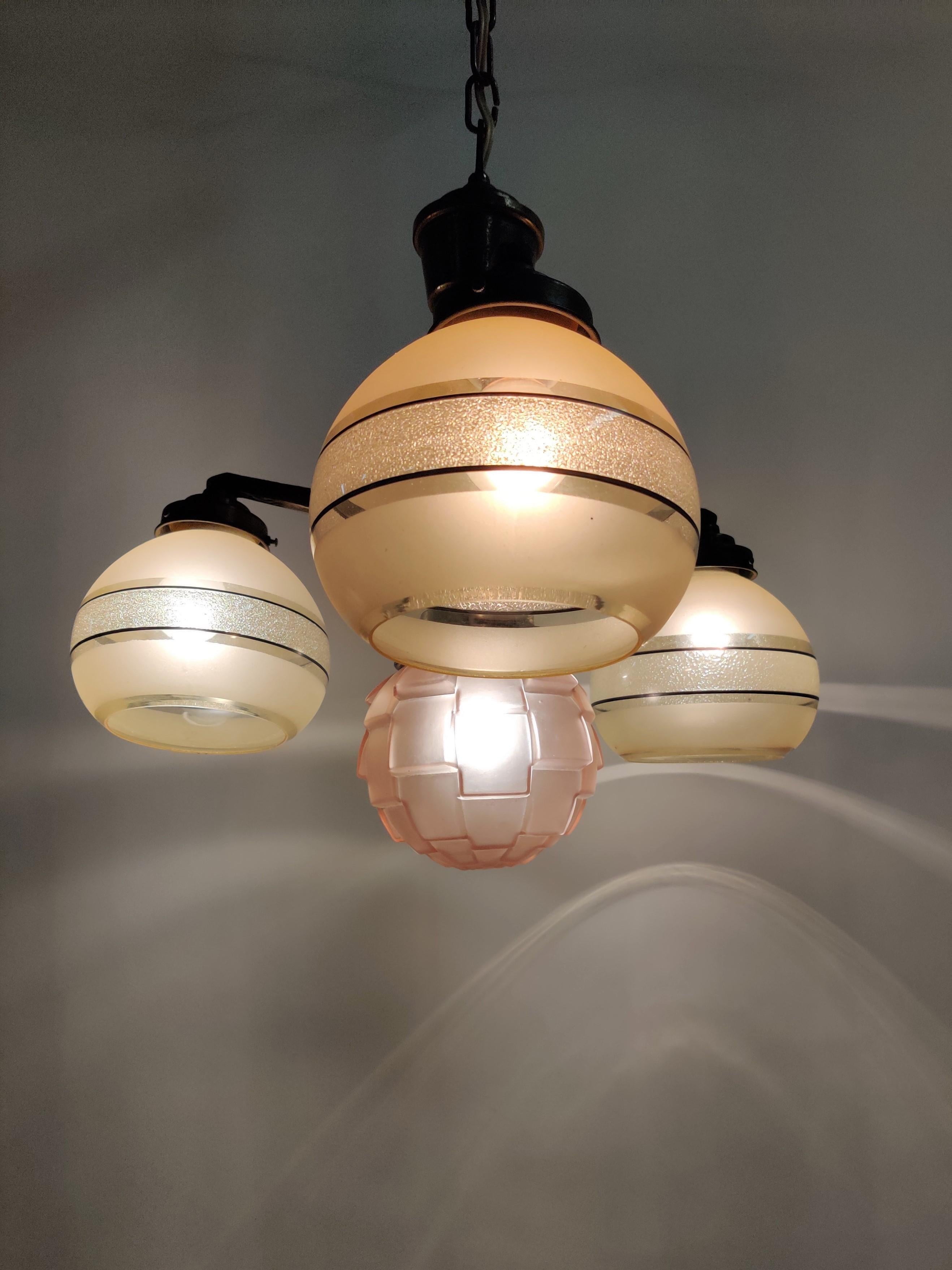 Art deco era chandelier with three amber colored decorated glass shades. 
This chandelier has a fourth special pink glass shade in the middel. 

This 4 lightpoint chandelier is made from patinated cast bronze.

Good original