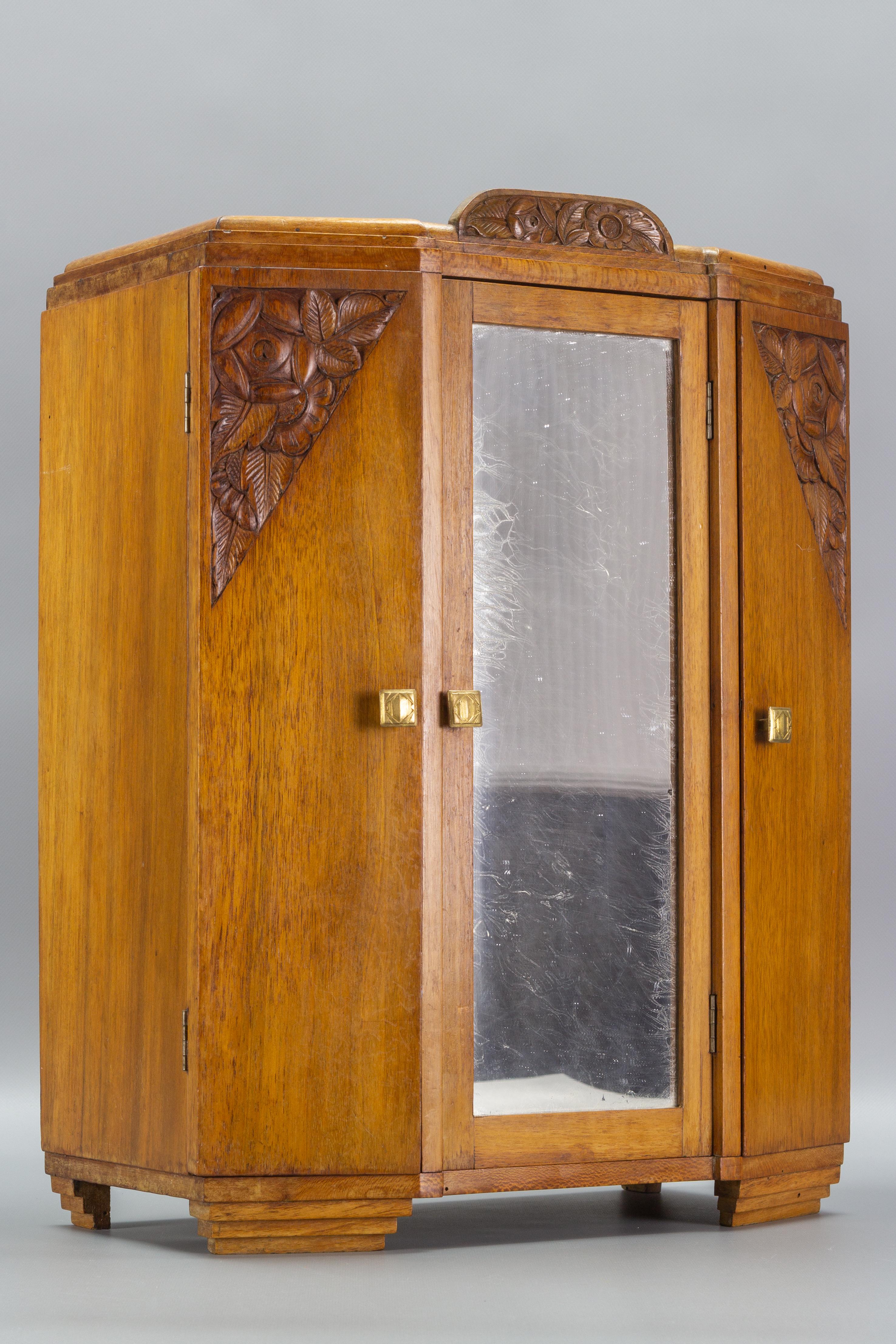 Unique and stunning French Art Deco three-door miniature armoire with mirror, brass handles, and hand-carved floral ornaments. Two removable shelves. 
Most probably it was a prototype of a large Art Deco cabinet designed and handcrafted by a master