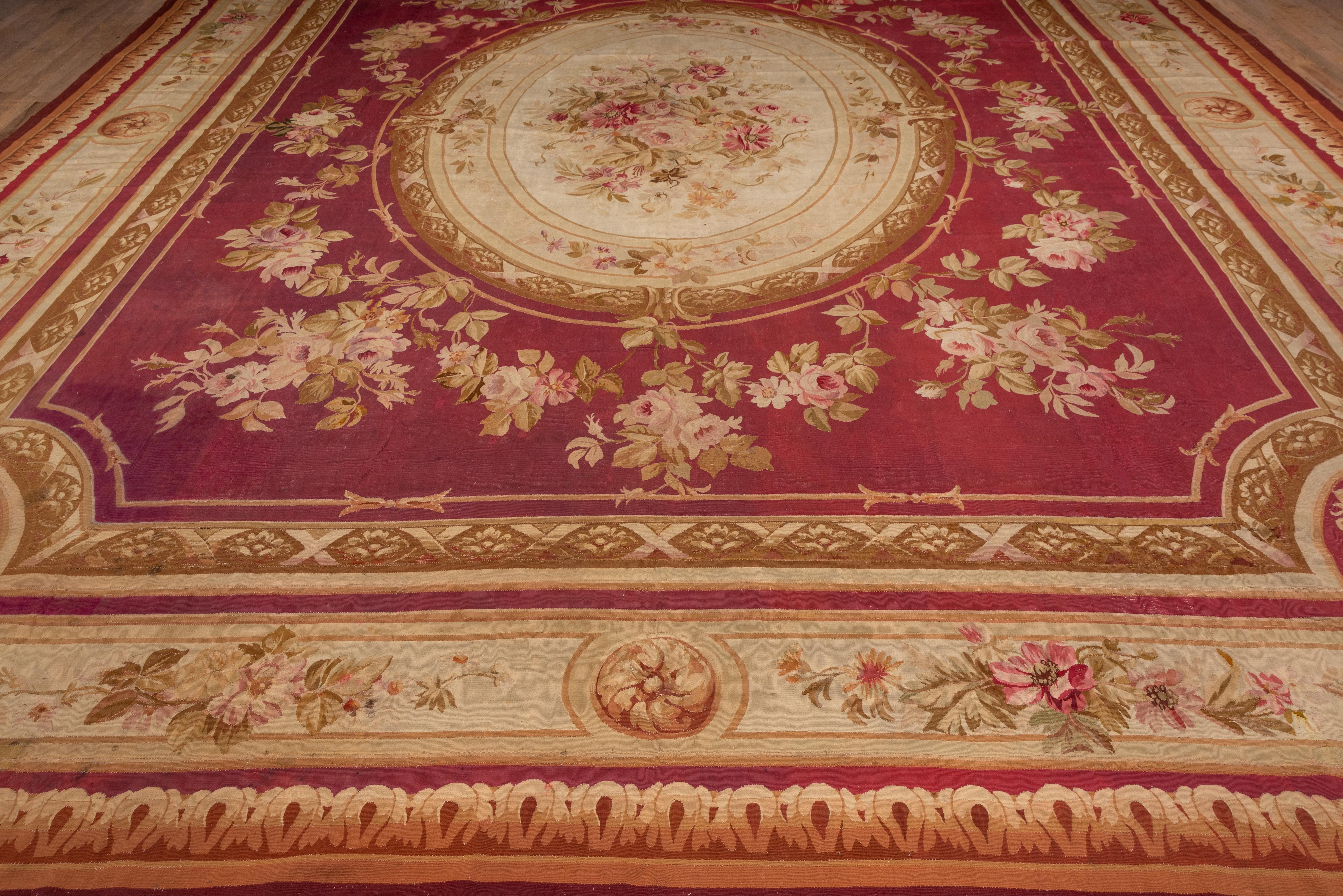 Fit for a Rothschild, this large antique French tapestry-woven carpet shows a large, layered oval medallion with an ivory central panel enclosing a detailed rose bouquet., all on a shaped red field with rose bouquets and swags, set with a cream main