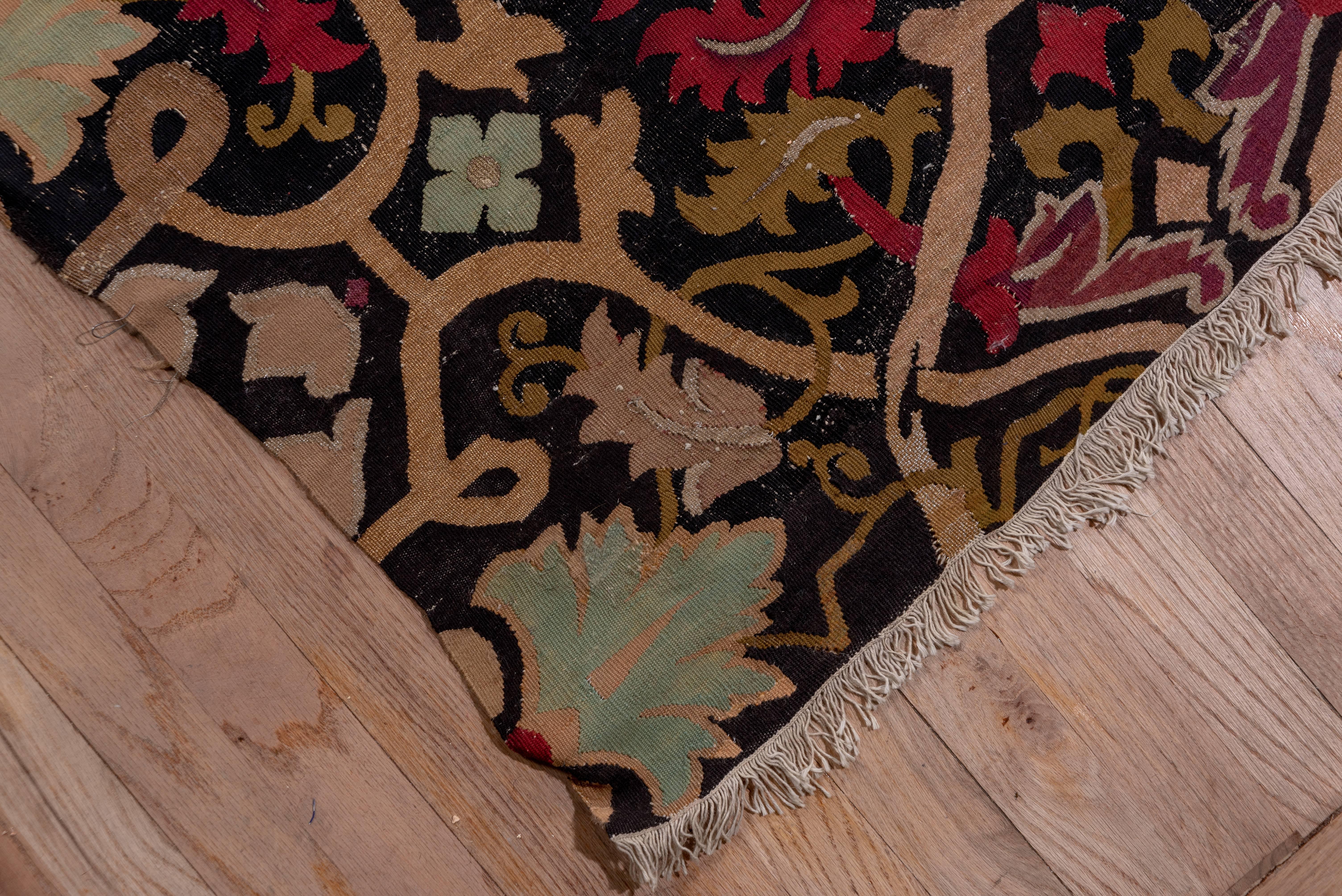 This border-less, black ground tapestry woven French pile-less carpet shows and allover pattern of rosette cruciforms, penetrating strap work vinery and a variety of acanthus leaves. There are accents in gold and silver tone metal thread as well as