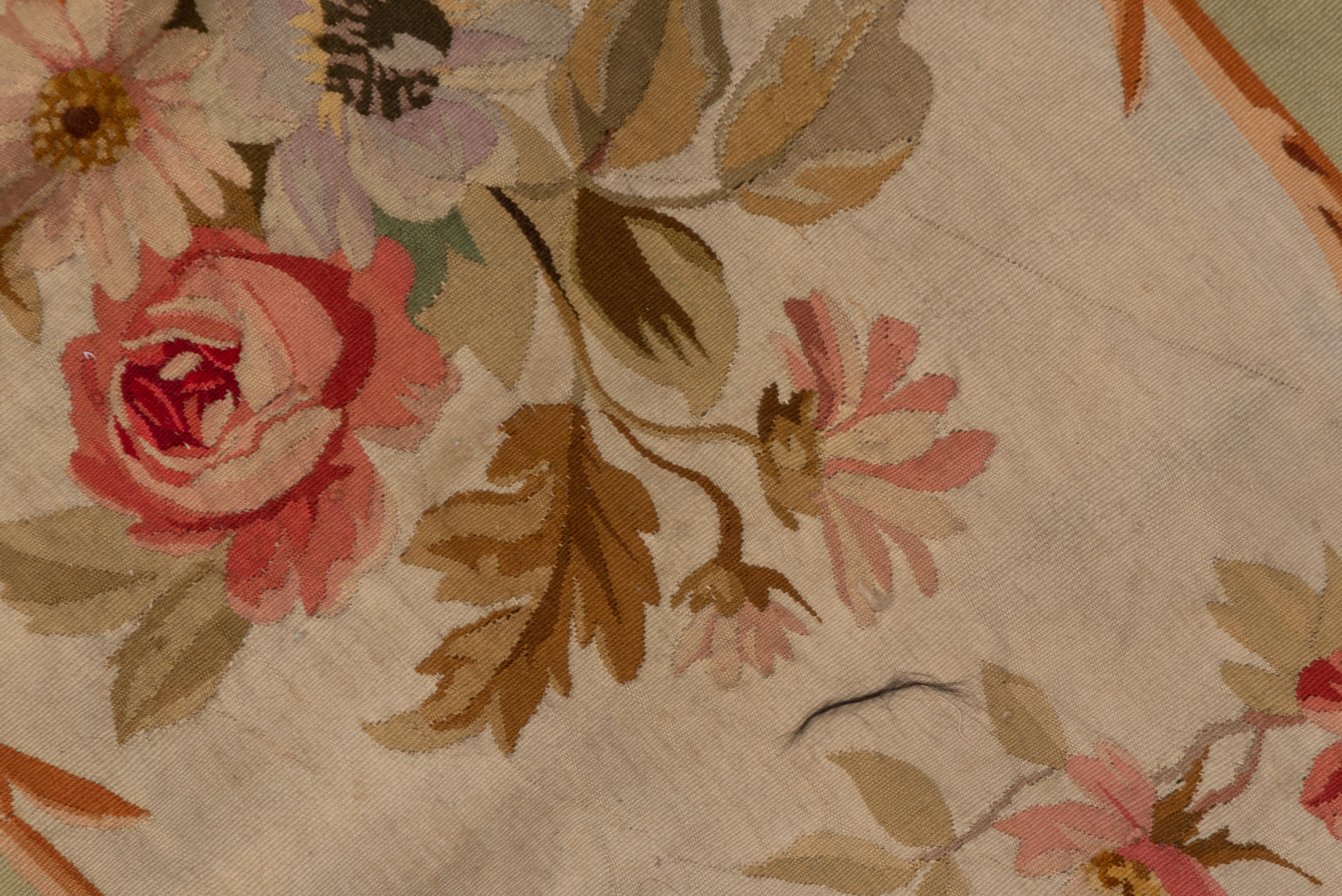 This slightly trapezoidal section of an antique French tapestry-woven carpet displays a broad ecru section with rose, carnation and sprays of other floral decor. Beige plain side areas. Generally good condition with a softer spring palette.