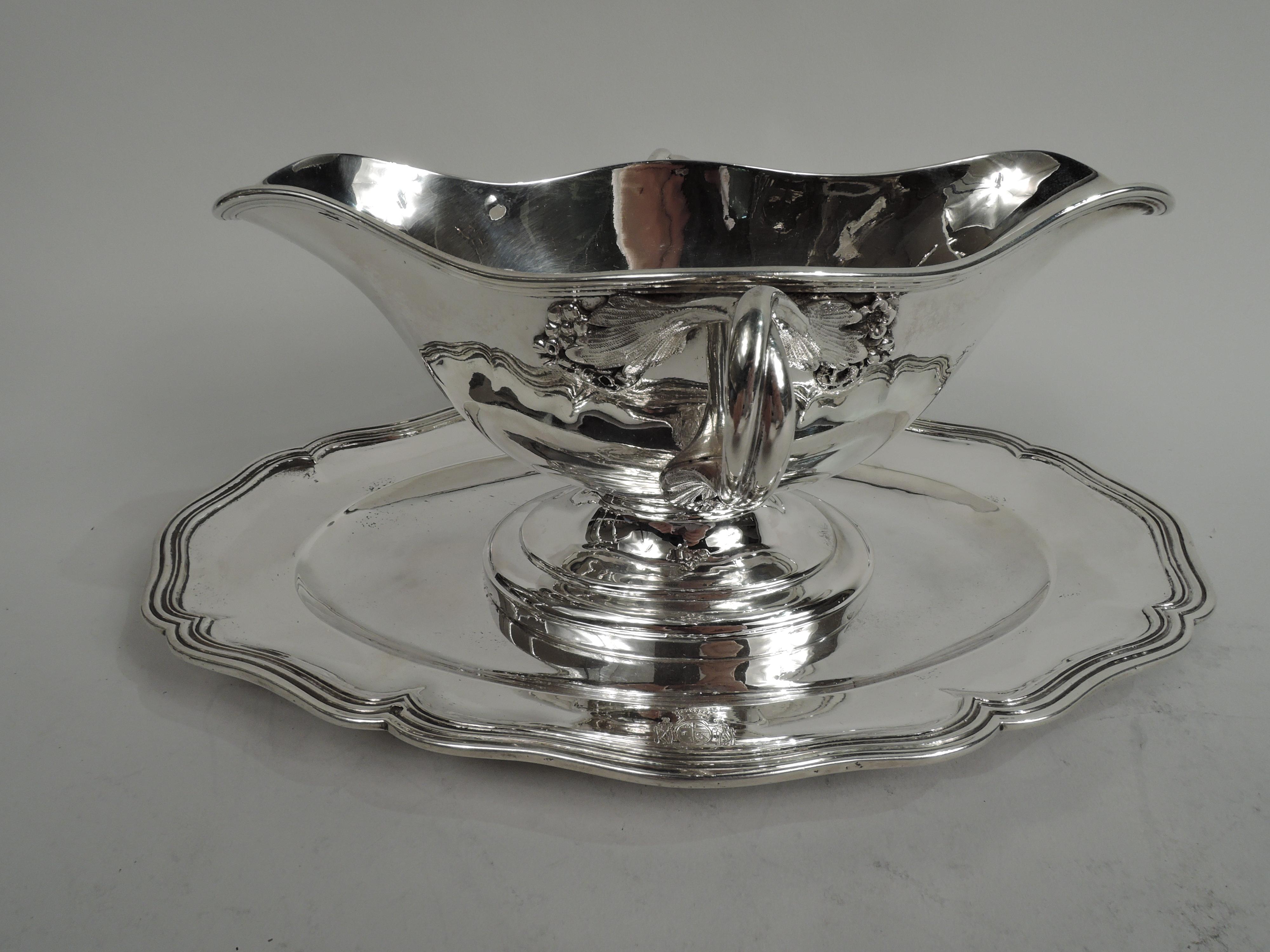 Turn-of-the-century French Belle Epoque Classical 950 silver gravy boat on stand. Oval bowl with curved sides and elongated end spouts. Side handles entwined and split-mounted with leaves and flowers. Stepped oval foot mounted to stand with oval