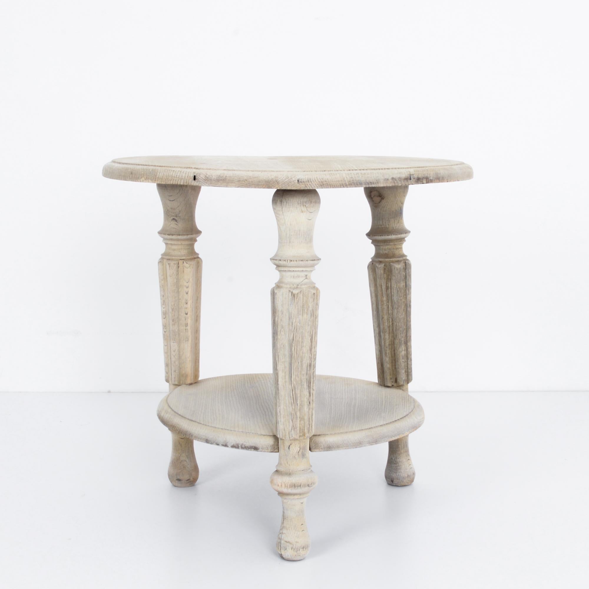 This bleached oak side table was made in France and brings the rustic and elegant charm of the countryside. It has a round tabletop with a molded edge and a lower shelf. Three fluted and tapered legs support the table. The smooth finish of the table