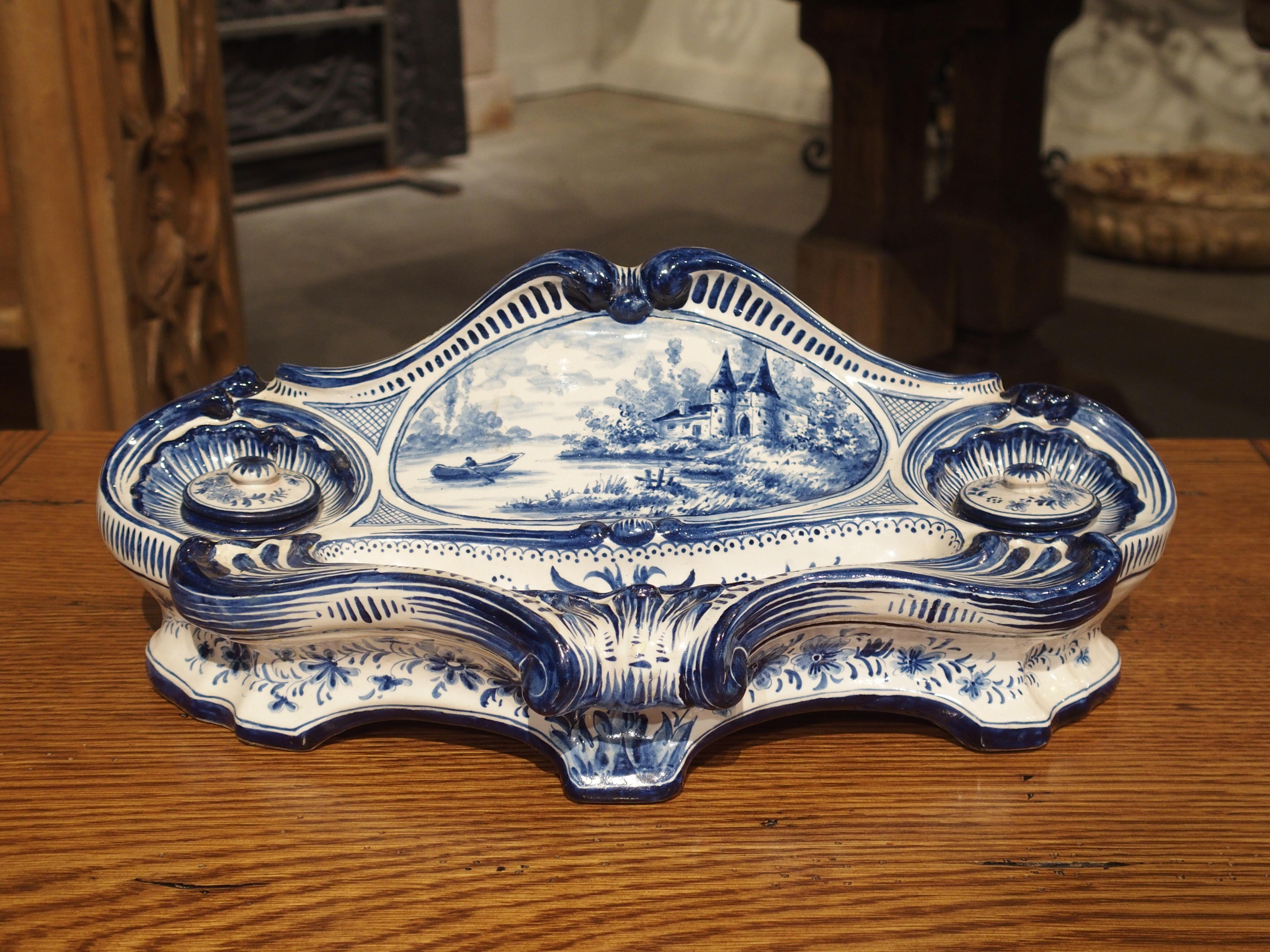 The charm of this French faience inkwell lies in the hand painted decor and its unique shape.

The central medallion depicts a person rowing a boat towards a chateau surrounded by woods on the edge of the body of water. Flanking this scene are two